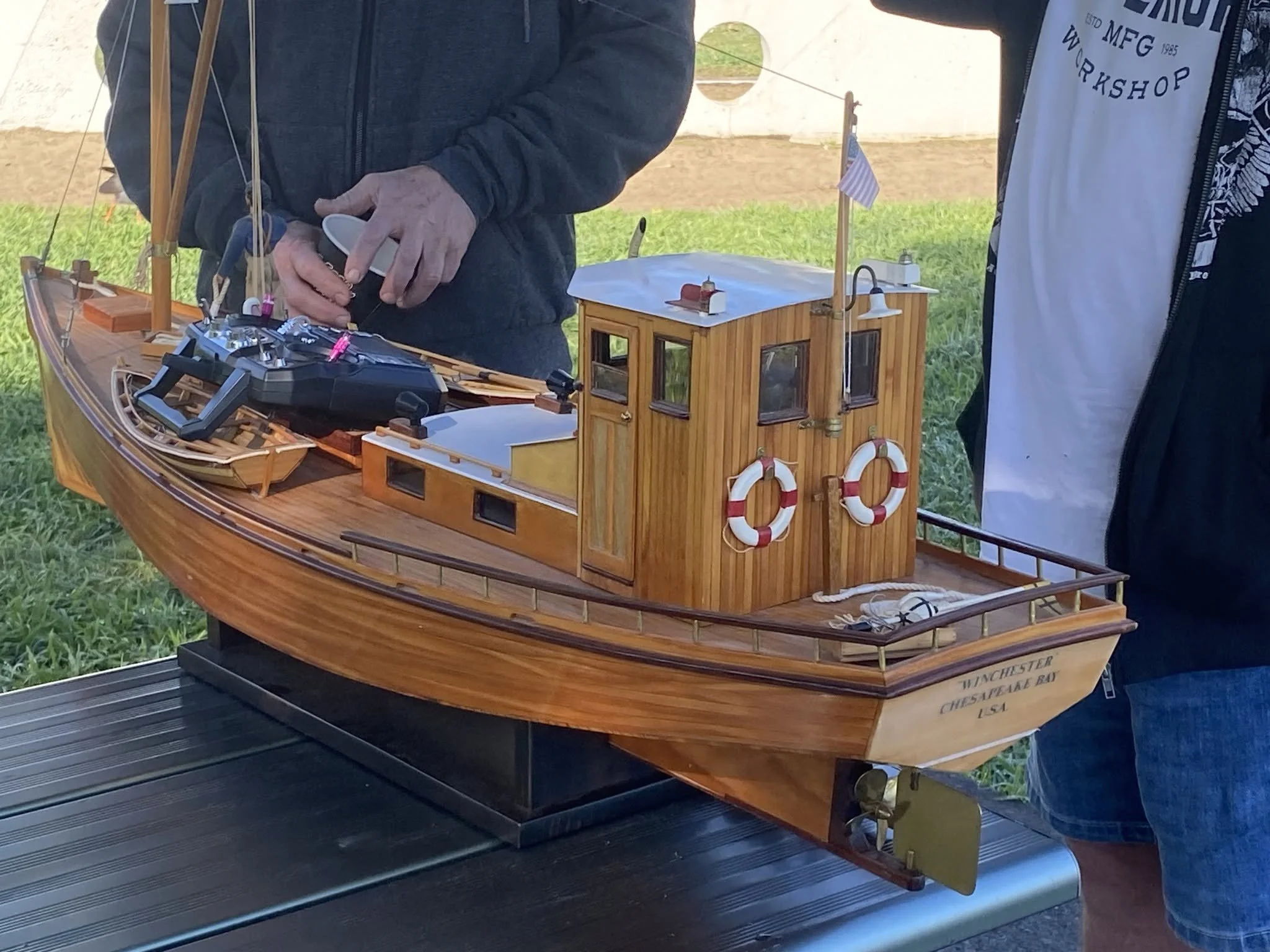 A detailed wooden model of a boat with small cabins and life preservers, with a person holding a remote control or device near the boat, on a black surface outdoors.