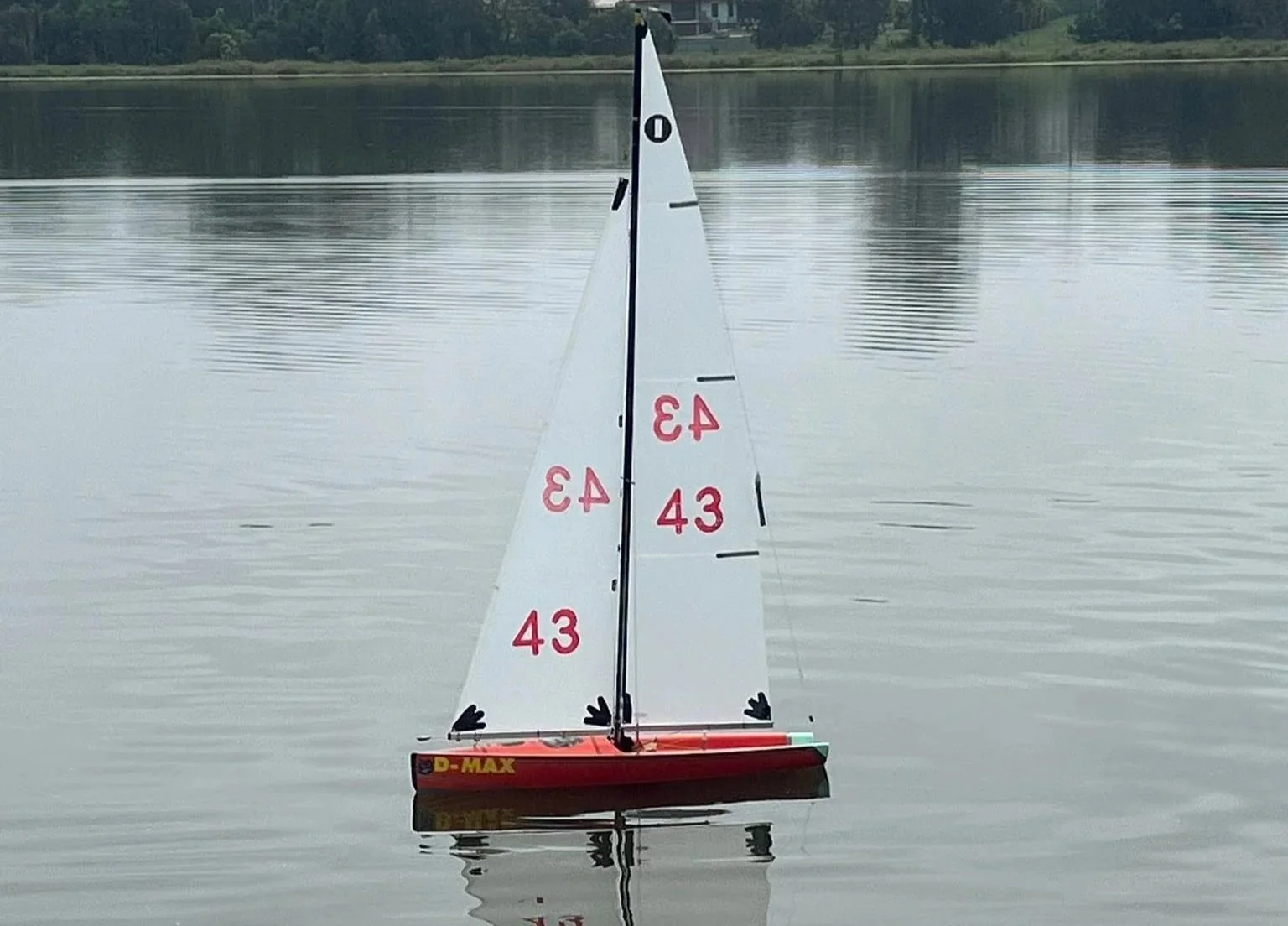 A remote-controlled toy sailboat with a red hull on calm water, with a background of trees and a cloudy sky.