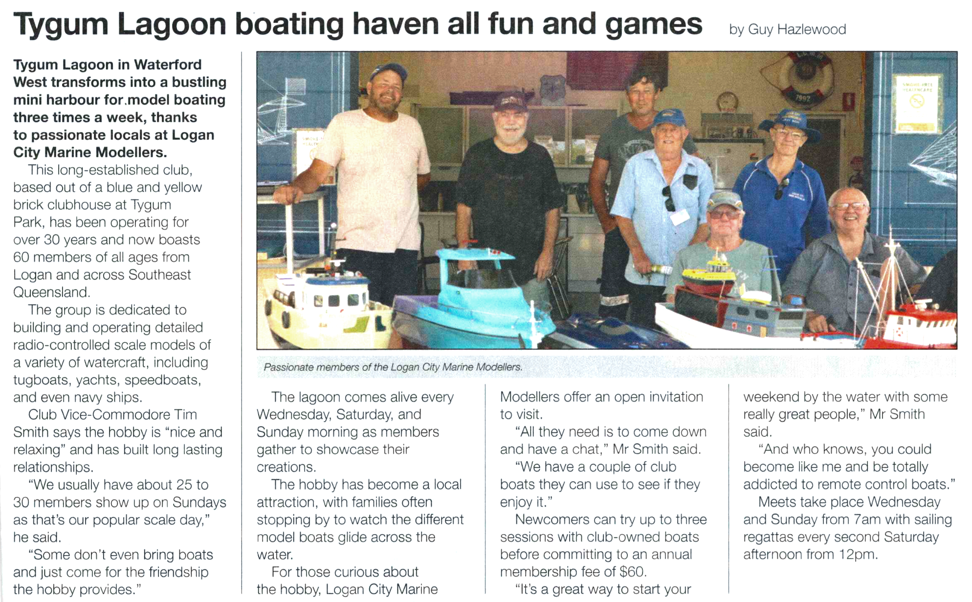 Photo of a group of people, members of the Logan City Marine Modellers, standing behind model boats on display inside a clubhouse or indoor area. The group is smiling, and the model boats include a tugboat, yacht, and speedboat.