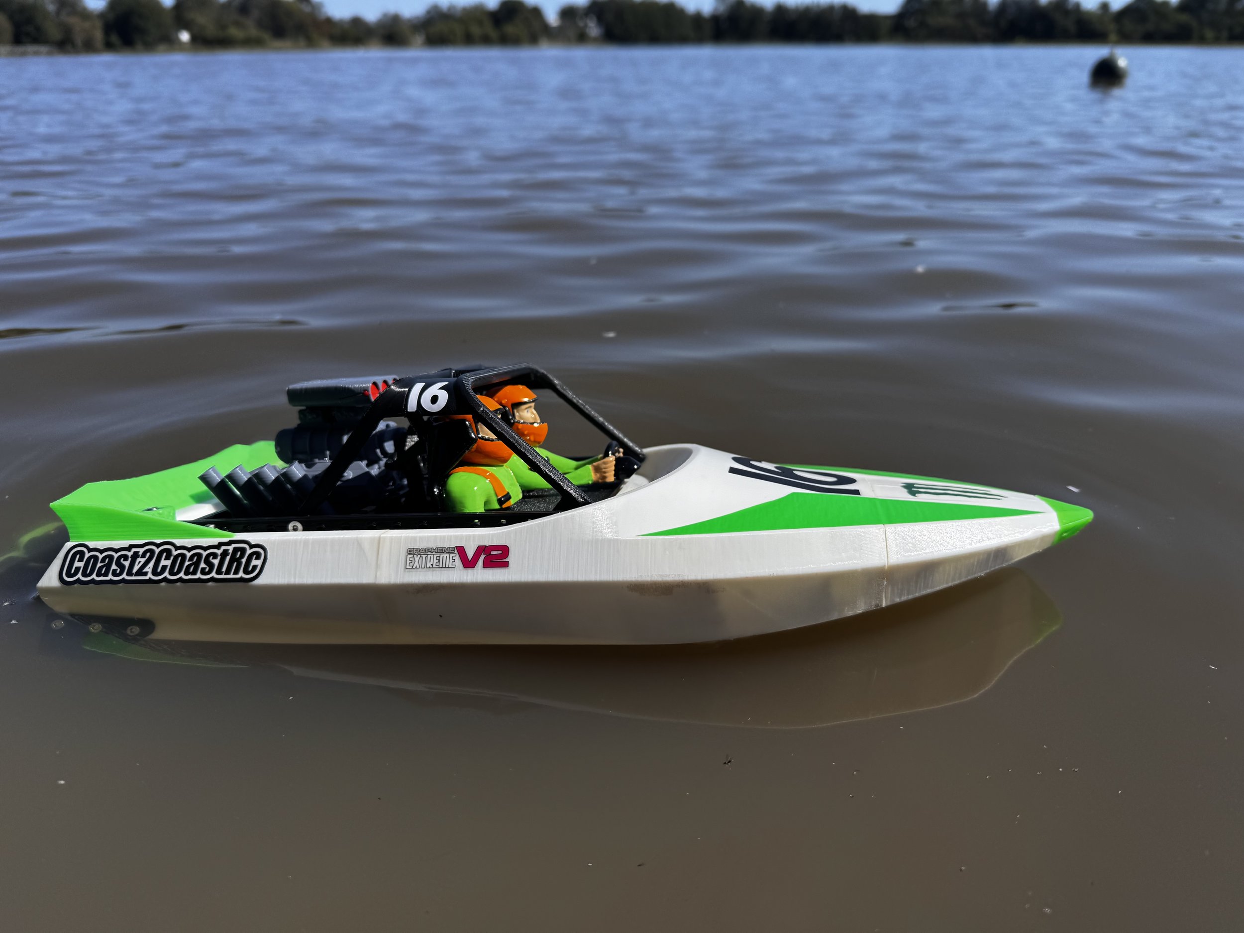 A remote-controlled toy boat floating on a lake with a dock in the background and trees on the shoreline
