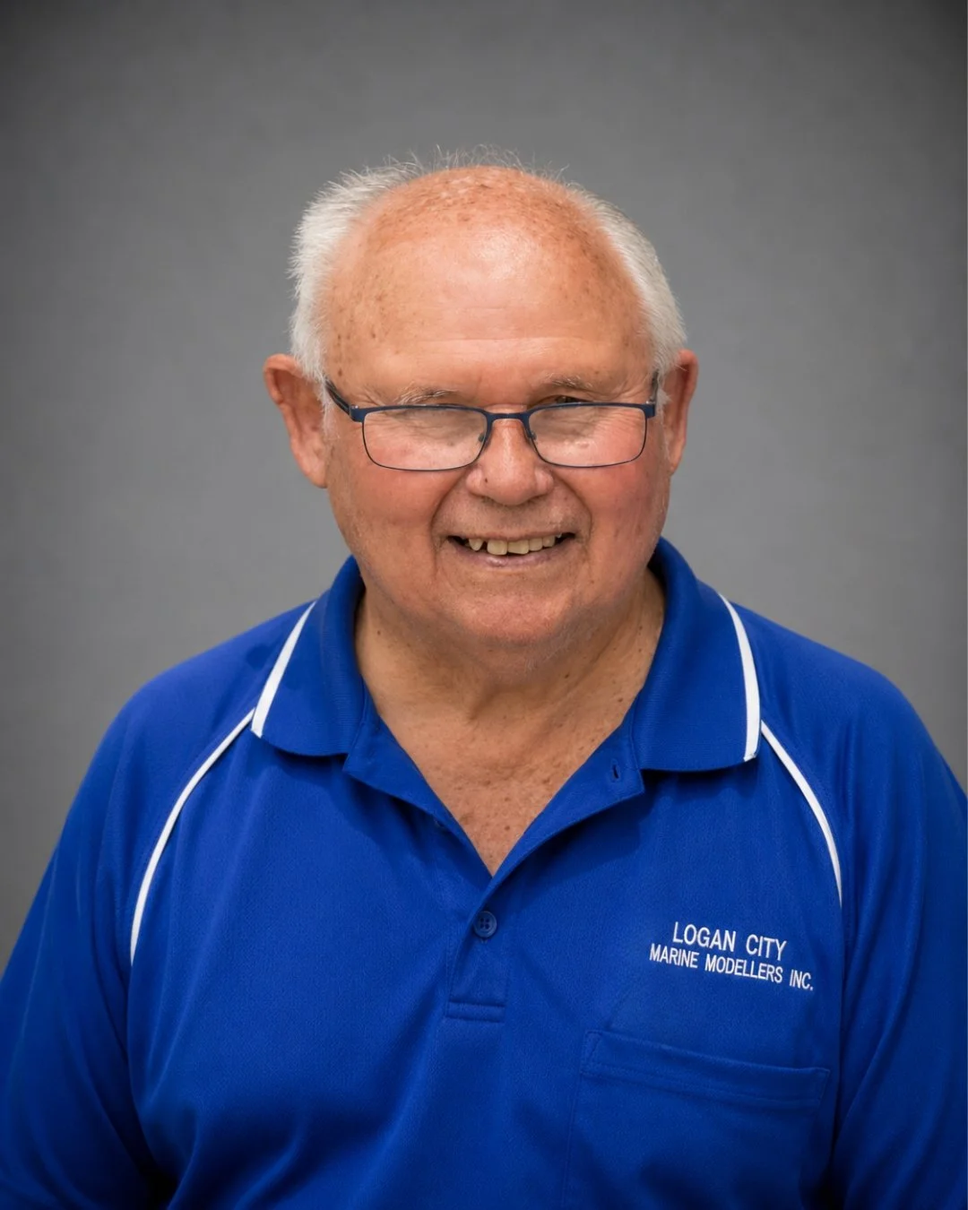Smiling elderly man with glasses wearing a blue polo shirt with white trim, with text that reads 'Logan City Marine Modellers Inc.', against a gray background.