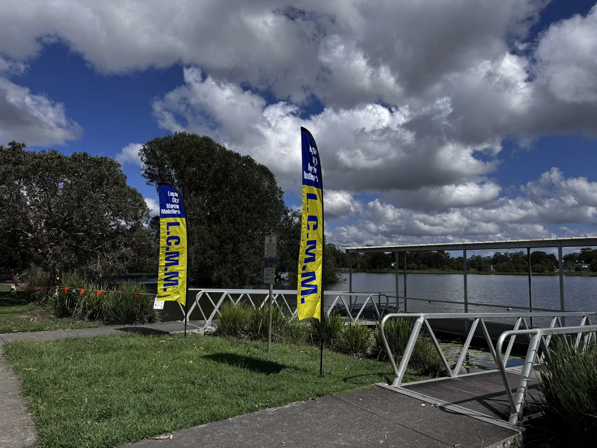 Two blue and yellow flags with text near a waterfront with trees and a cloudy sky.