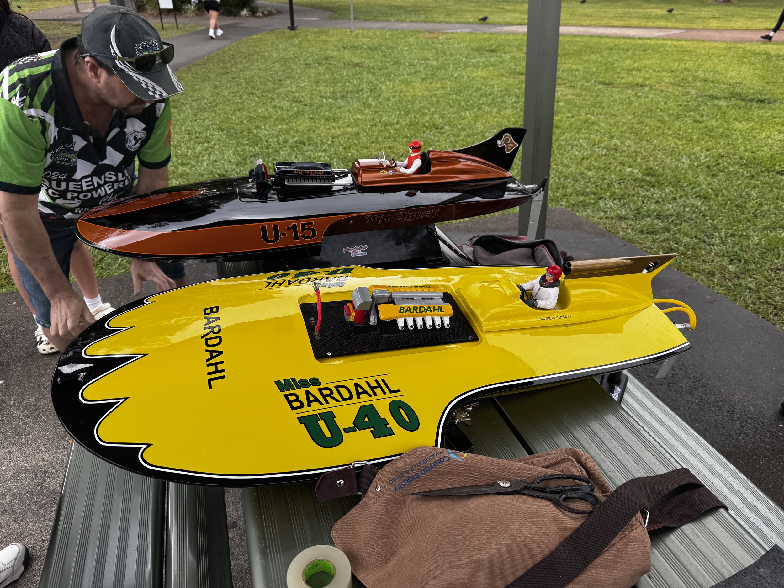 Two remote-controlled cars resembling racing cars, one yellow and one black and orange, are placed on a table outside. The yellow car has 'Miss Bardahl' and 'U-40' written on it, while the black and orange car features 'U-15' and 'High Gear' logos. A