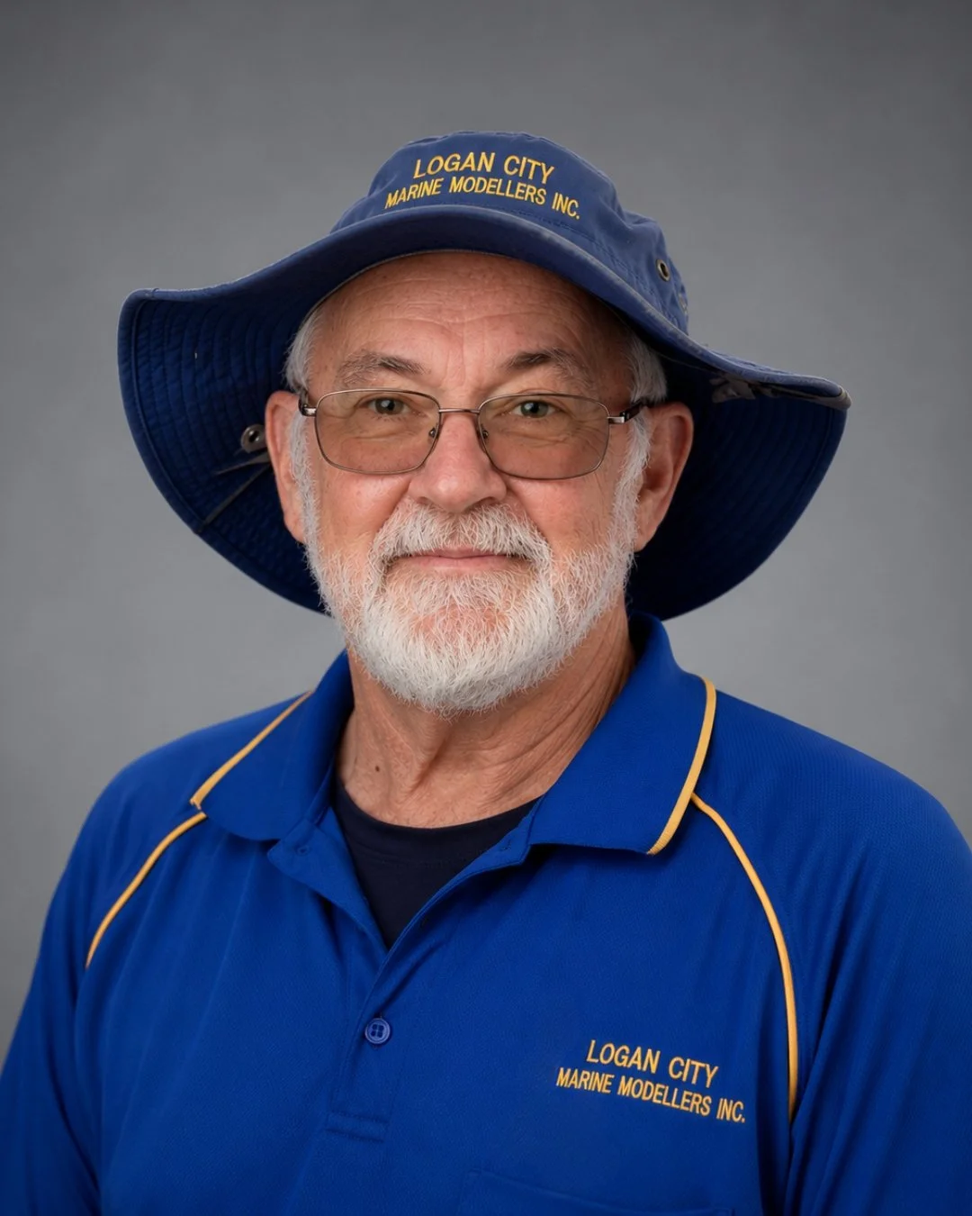 An elderly man with a white beard and glasses wearing a blue Logan City Marine Modellers Inc. polo shirt and a wide-brimmed blue hat, posing against a gray background.