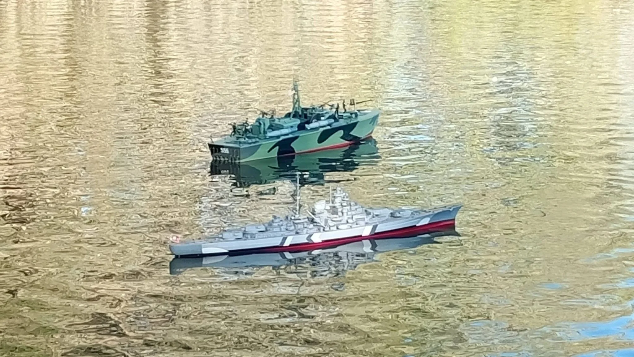 Two model ships floating on a water surface with a golden hue. One ship has a camouflage pattern, and the other is gray with maritime markings.