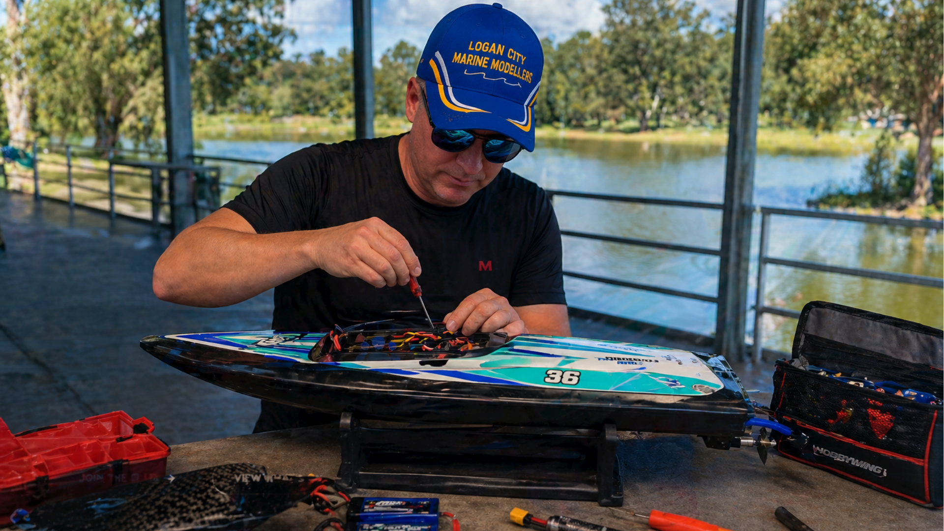 A man wearing a blue cap with yellow lettering that reads 'Logan City Marine Modellers' and sunglasses is working on a remote-controlled boat at a table outdoors near a body of water. He is adjusting wiring on the boat with various tools and equipment spread out on the table.