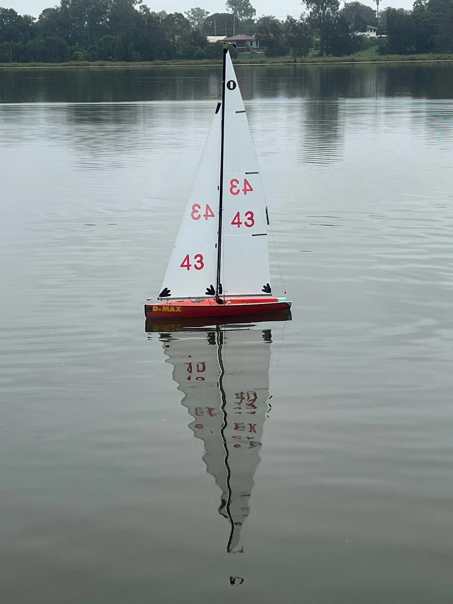 A model sailboat floating on a calm body of water, with a reflection visible on the water's surface. In the background, there are trees, a few houses, and an overcast sky.