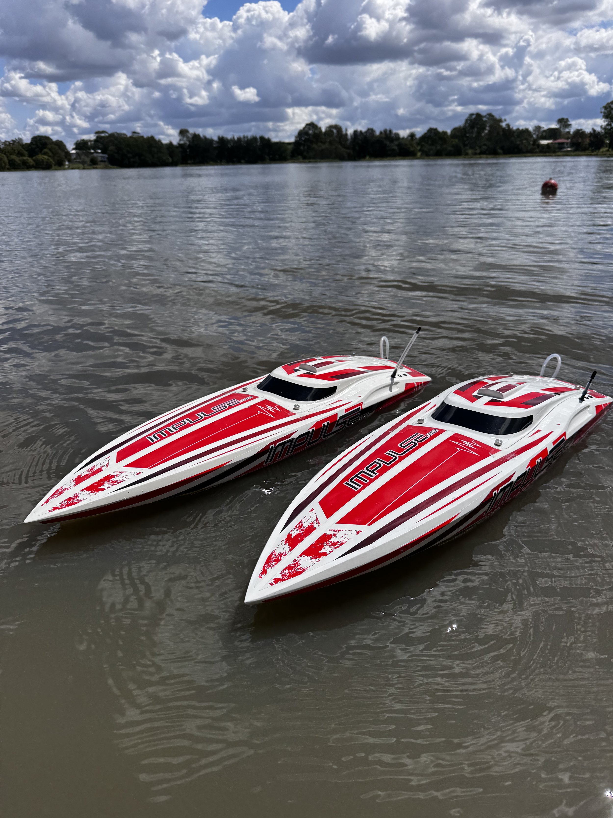 Two remote-controlled speedboats with red and white designs floating on a lake, with trees and houses in the background under a partly cloudy sky.