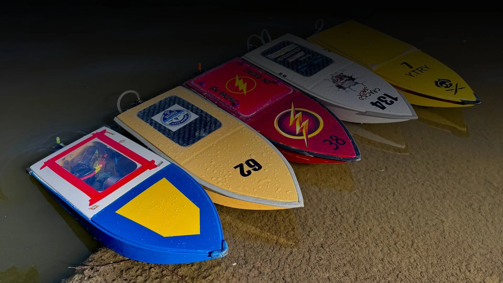 Four colorful paddleboards with various designs and logos are lined up on the sandy beach near the water.