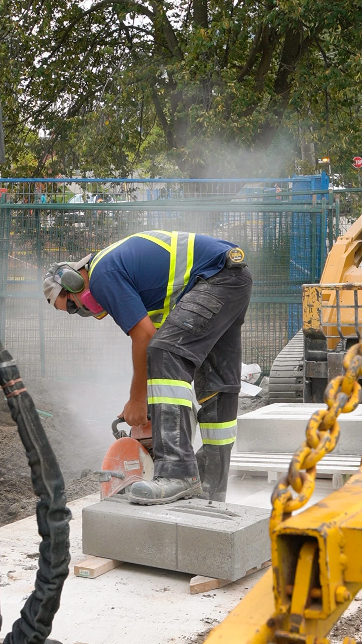 A construction worker wearing a safety vest, headphones, and a cap is cutting a concrete block with a power saw on a construction site. Dust is visible in the air.