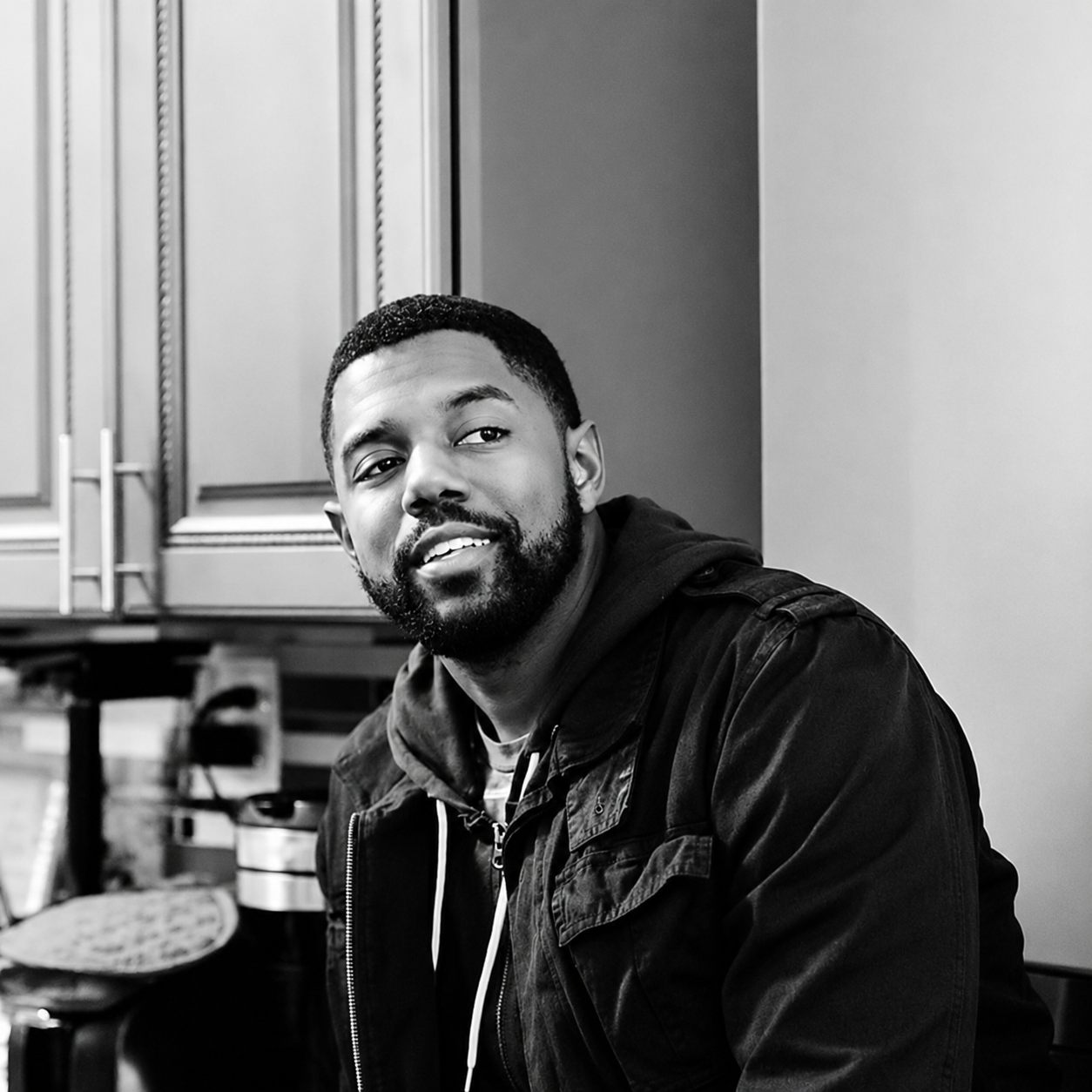 A young man with a beard smiling in a kitchen, wearing a dark jacket and hoodie, with kitchen cabinets in the background.