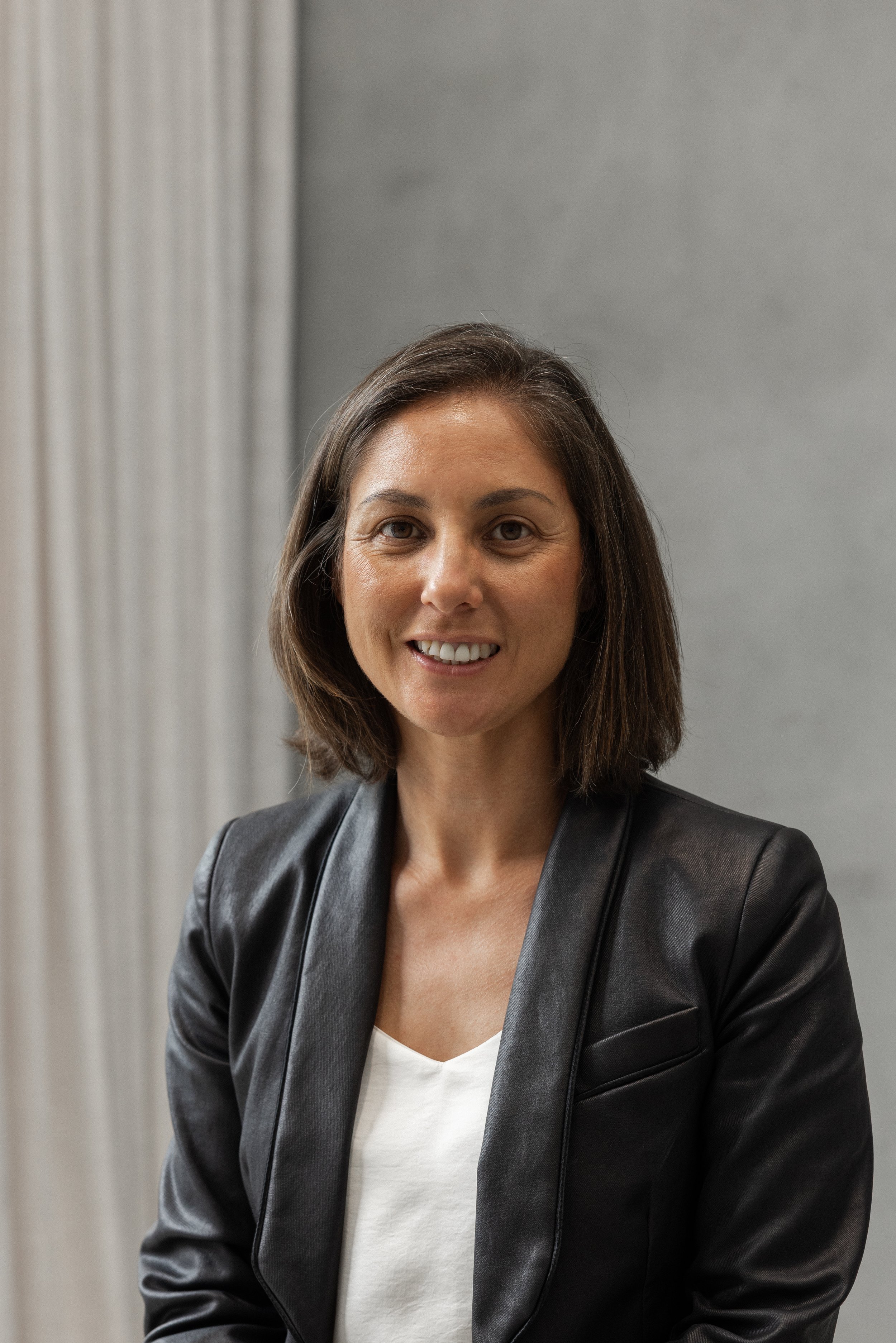 A woman with shoulder-length brown hair wearing a black leather jacket over a white shirt, standing indoors in front of a gray wall and white curtains.