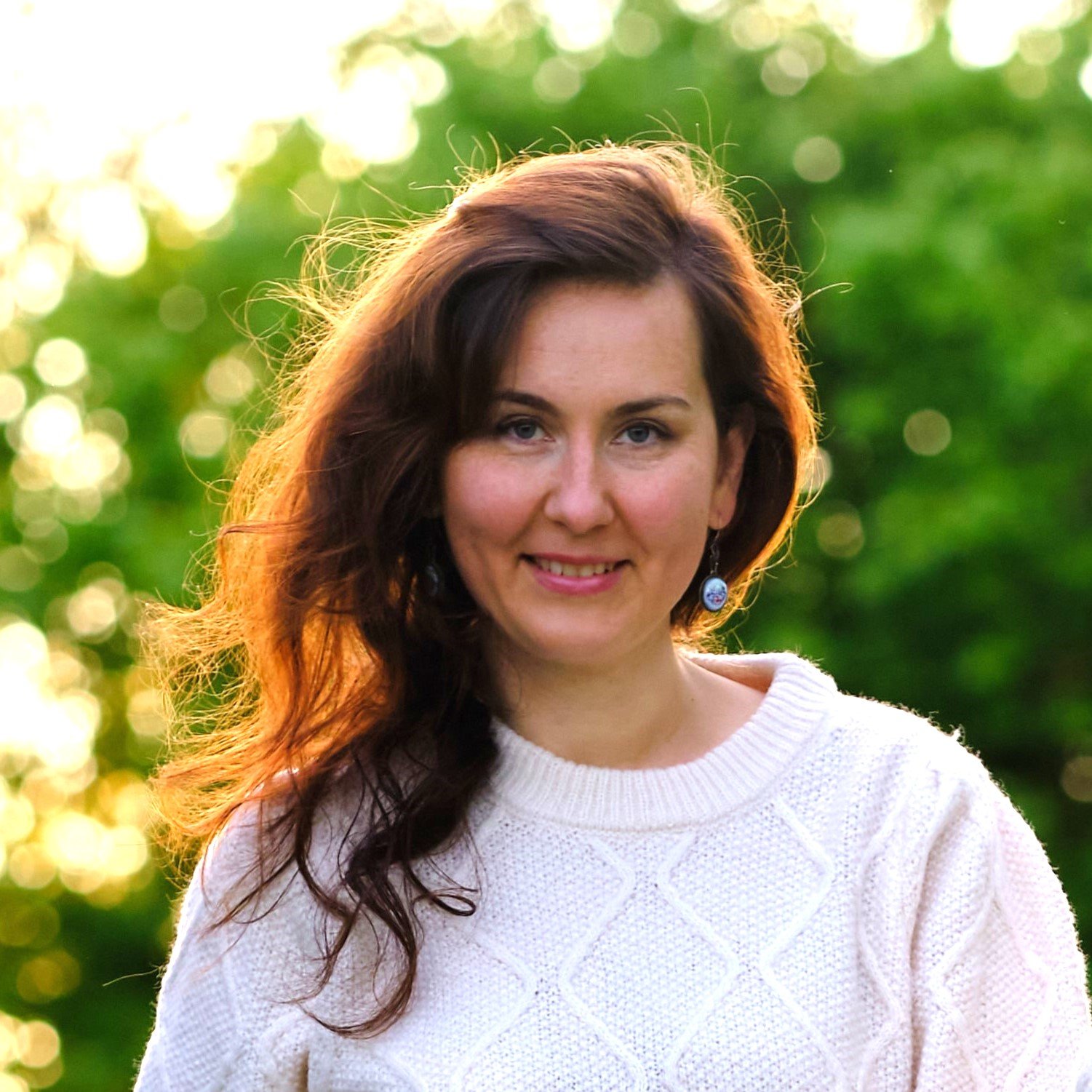 A woman with brown, wavy hair and blue eyes, smiling in a white knitted sweater outdoors with green trees and sunlight in the background.