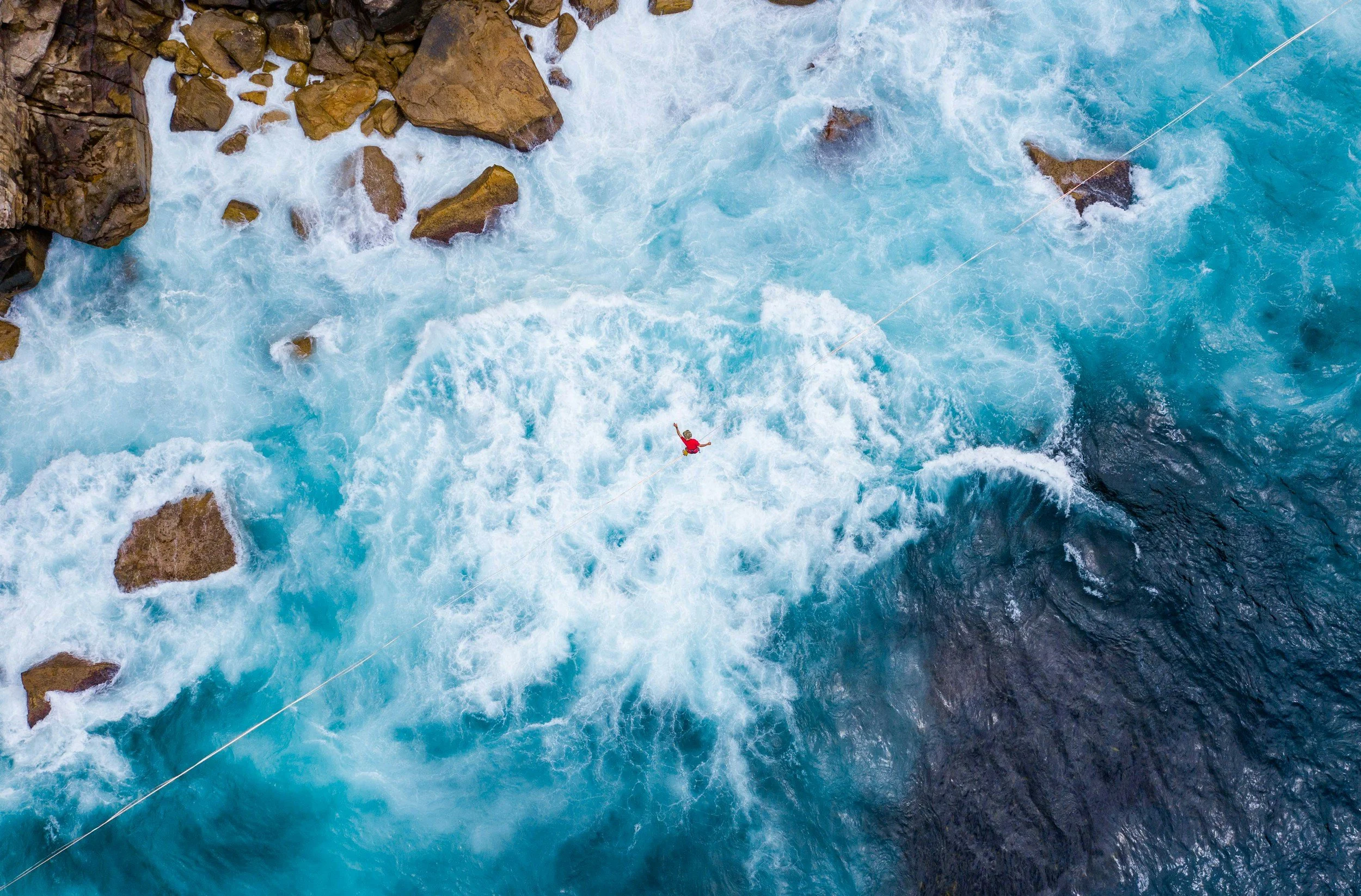An aerial view of a person in a red jacket and helmet white water rafting on a fast-moving, blue river surrounded by rocks.