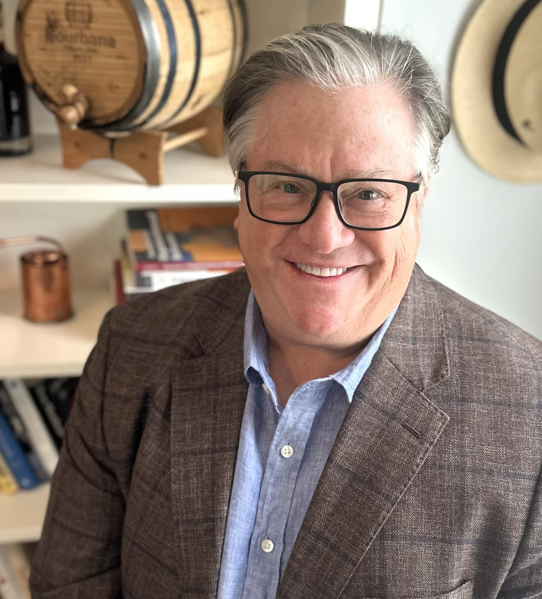 Close-up of a smiling man with gray hair and black glasses, wearing a brown blazer and light blue shirt, standing in a room with books and a wooden barrel in the background.