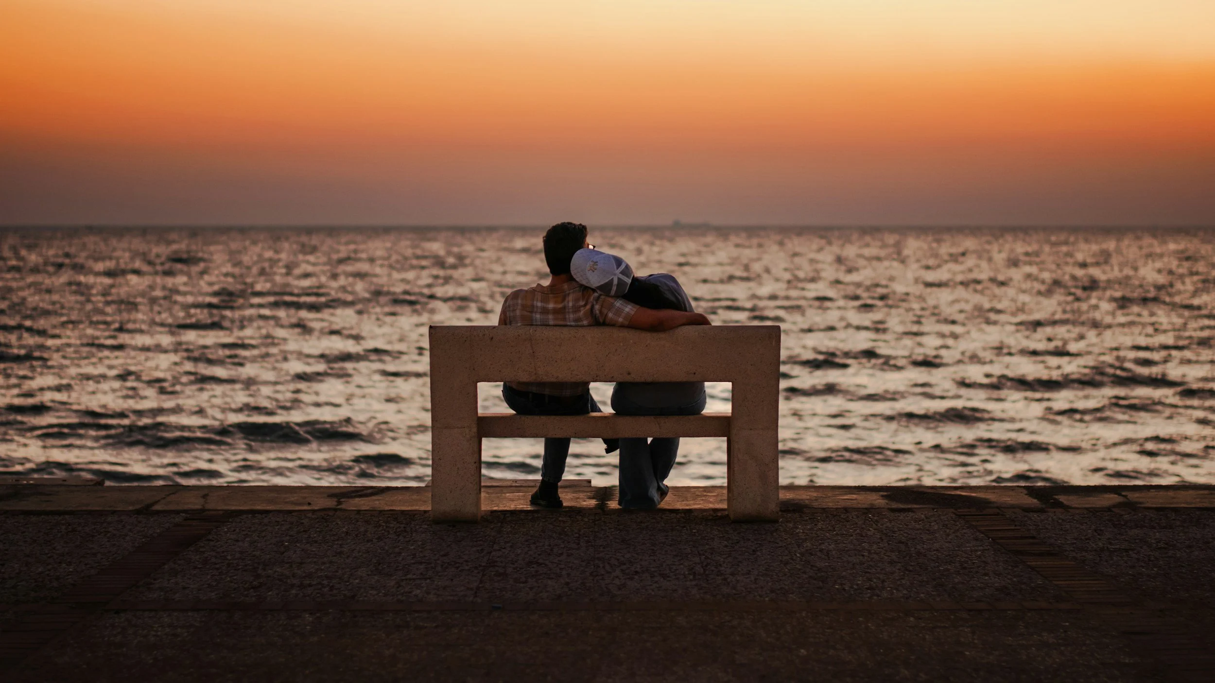 A couple sitting on a bench at the beach during sunset, with the ocean in the background and a soft pink and orange sky.