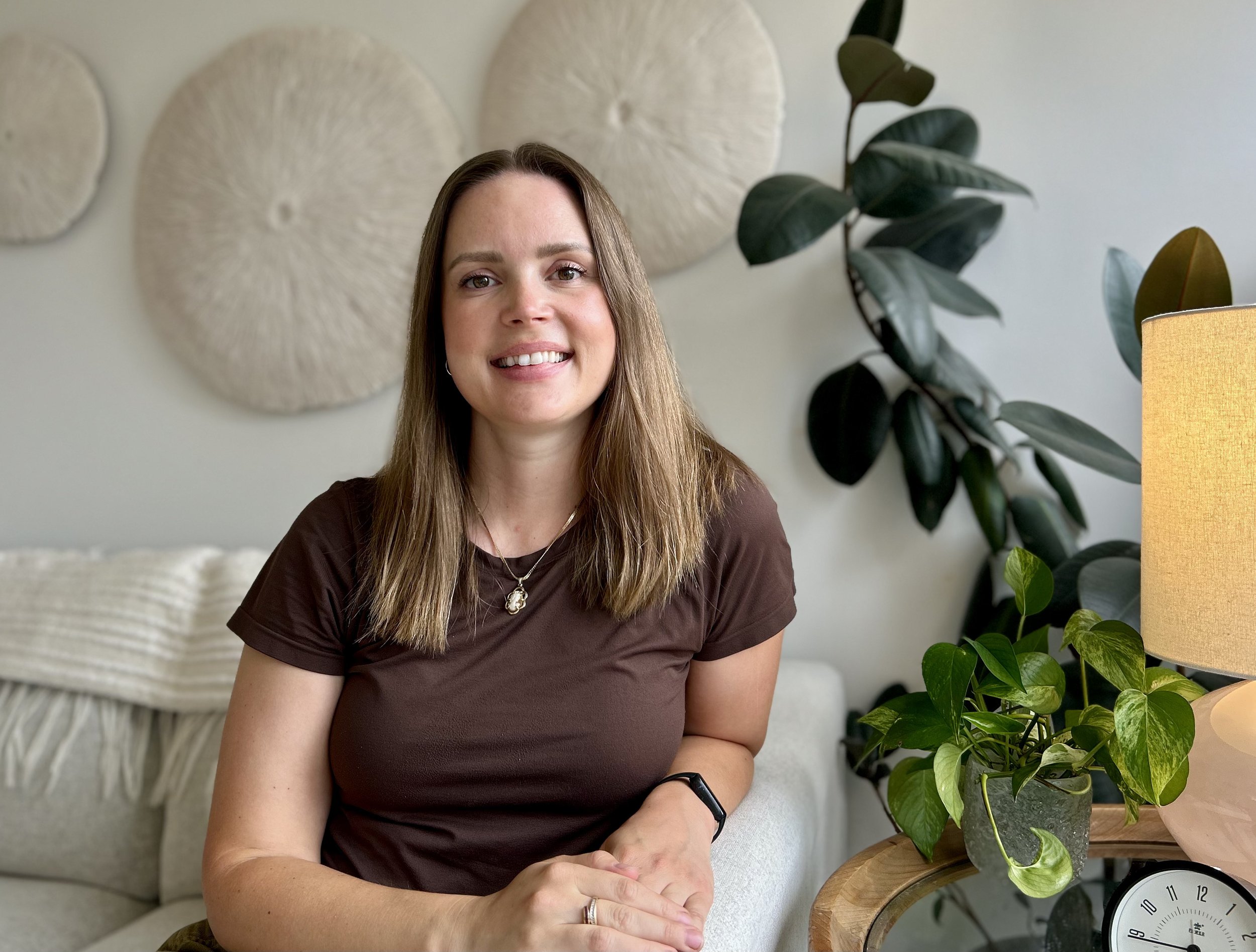 A woman with long brown hair, wearing a brown shirt and a necklace with a pendant, sitting in a room decorated with wall art and plants, smiling at the camera.