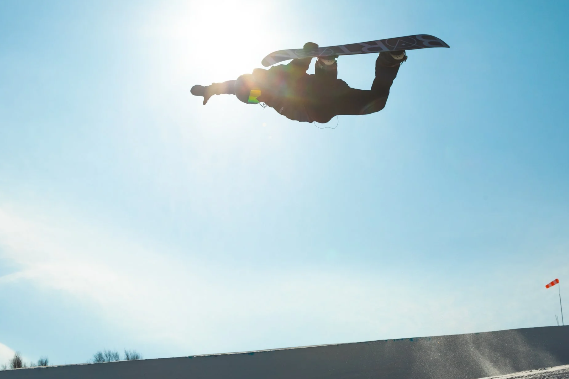 A person standing on a raised platform outdoors, holding a snowboard, with the sun shining behind them against a clear blue sky.