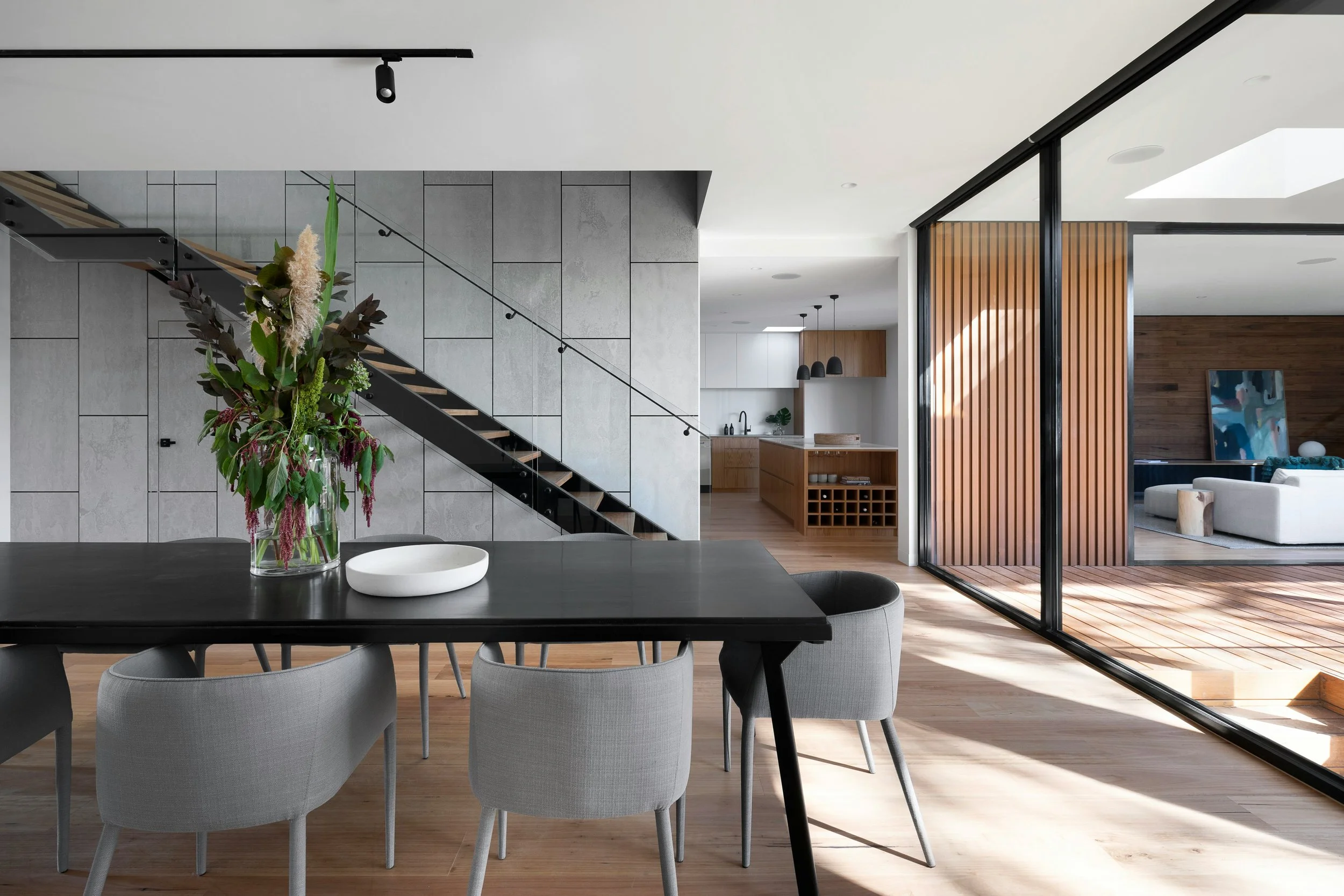 Modern open-concept living space with a dining area, staircase, and kitchen, featuring large windows, wooden accents, and a decorative flower arrangement on the table.