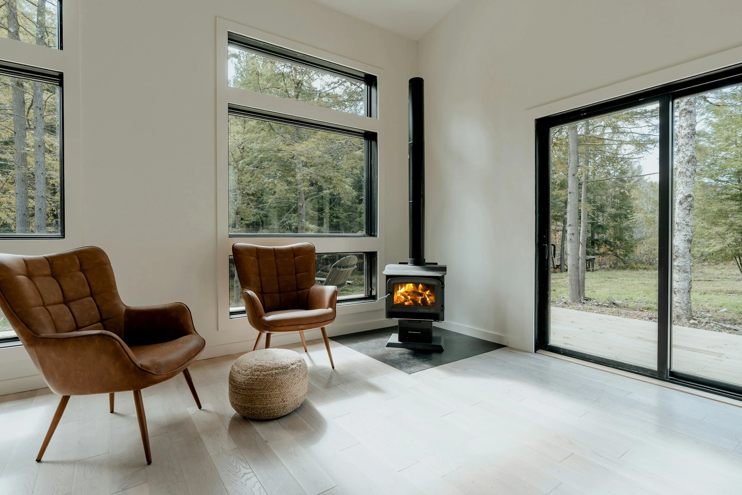 A living room with large windows and a sliding glass door, a wood stove with a fire burning, two brown leather chairs, a woven pouf, and a view of trees outside.