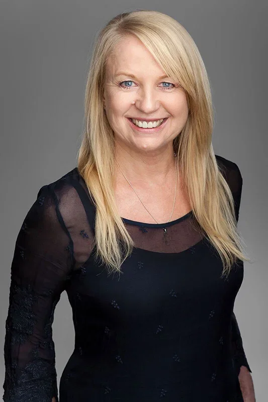A woman with long blonde hair, blue eyes, and a bright smile, wearing a black sheer top with lace details and a silver necklace, standing against a gray background.