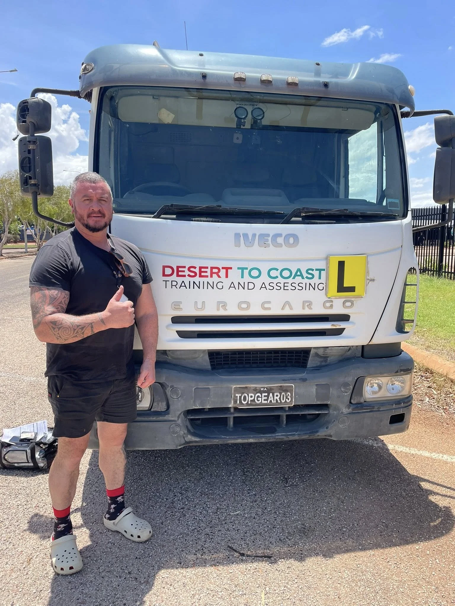 A man with tattoos on his left arm, wearing a black T-shirt, black shorts, red and black socks, and beige Crocs, giving a thumbs up and standing in front of a large white truck with the words "Desert to Coast Training and Assessing" and a yellow "L" sign on it. The truck has the brand name "IVECO" on the front and a license plate reading "TOPGEAR03." The background shows a partly cloudy sky and some trees.