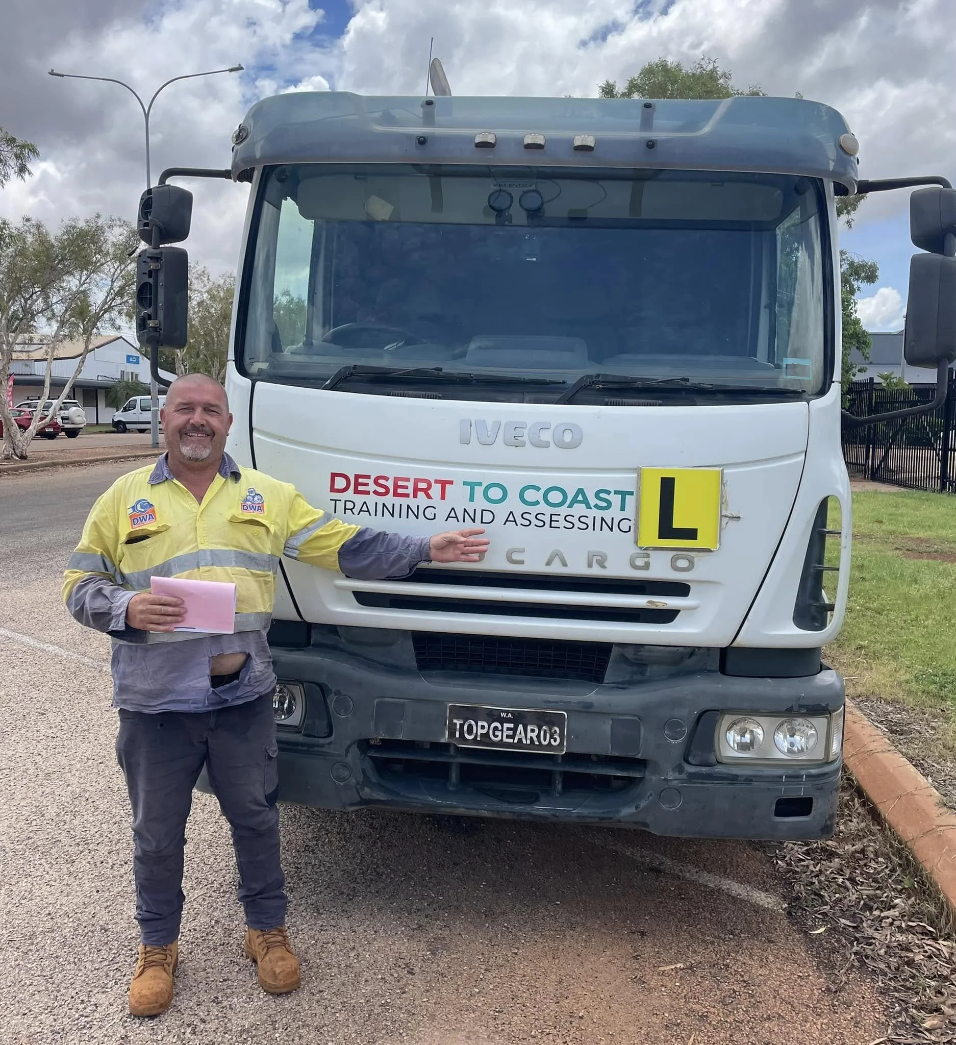 A man standing in front of a large white Iveco truck with a banner reading "Desert to Coast Training and Assessing" on the front. The man is smiling, wearing a yellow safety vest, brown boots, and holding pink papers.