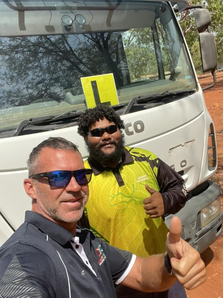 Two men taking a selfie in front of a white truck on a dirt road, both wearing sunglasses and smiling, one giving a thumbs up and the other holding a camera.