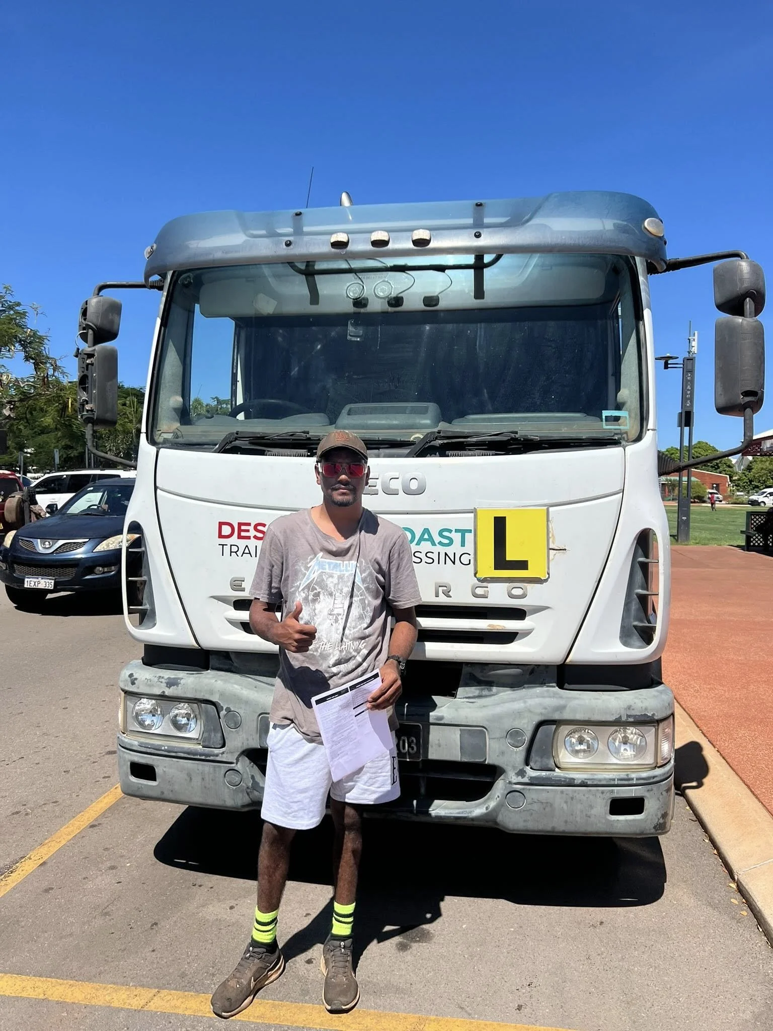 A young man stands in front of a large white truck with an 'L' sign, holding a document in one hand and giving a thumbs-up with the other. The man is wearing a gray Metallica T-shirt, white shorts, brown shoes, and neon yellow and black striped socks. The truck is parked in a lot on a sunny day, with several cars and trees visible in the background.
