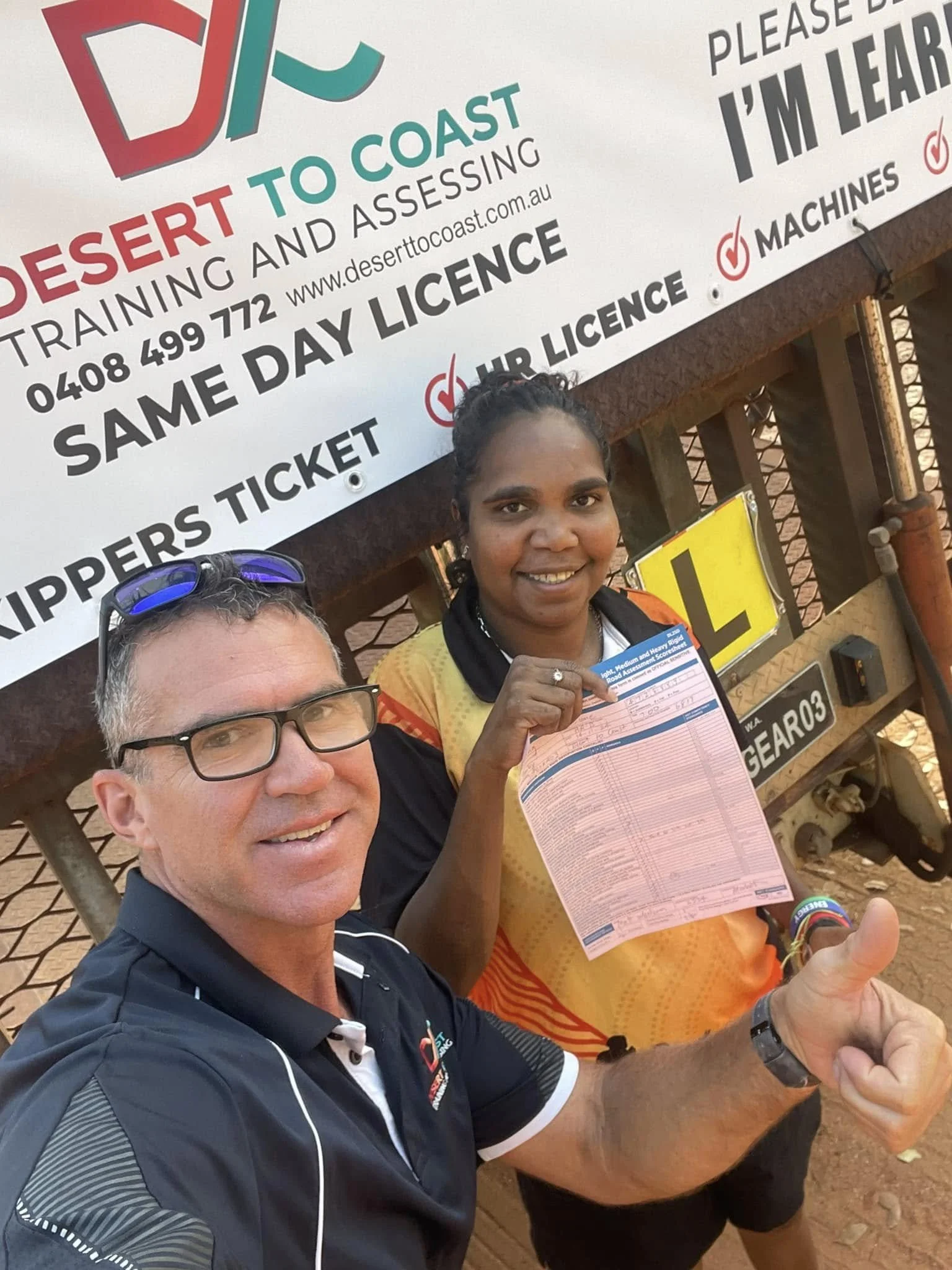 Two smiling people, one man and one woman, taking a selfie at a desert training and assessment event, with a large sign overhead. The woman is holding a pink paper, and both are giving a thumbs-up.