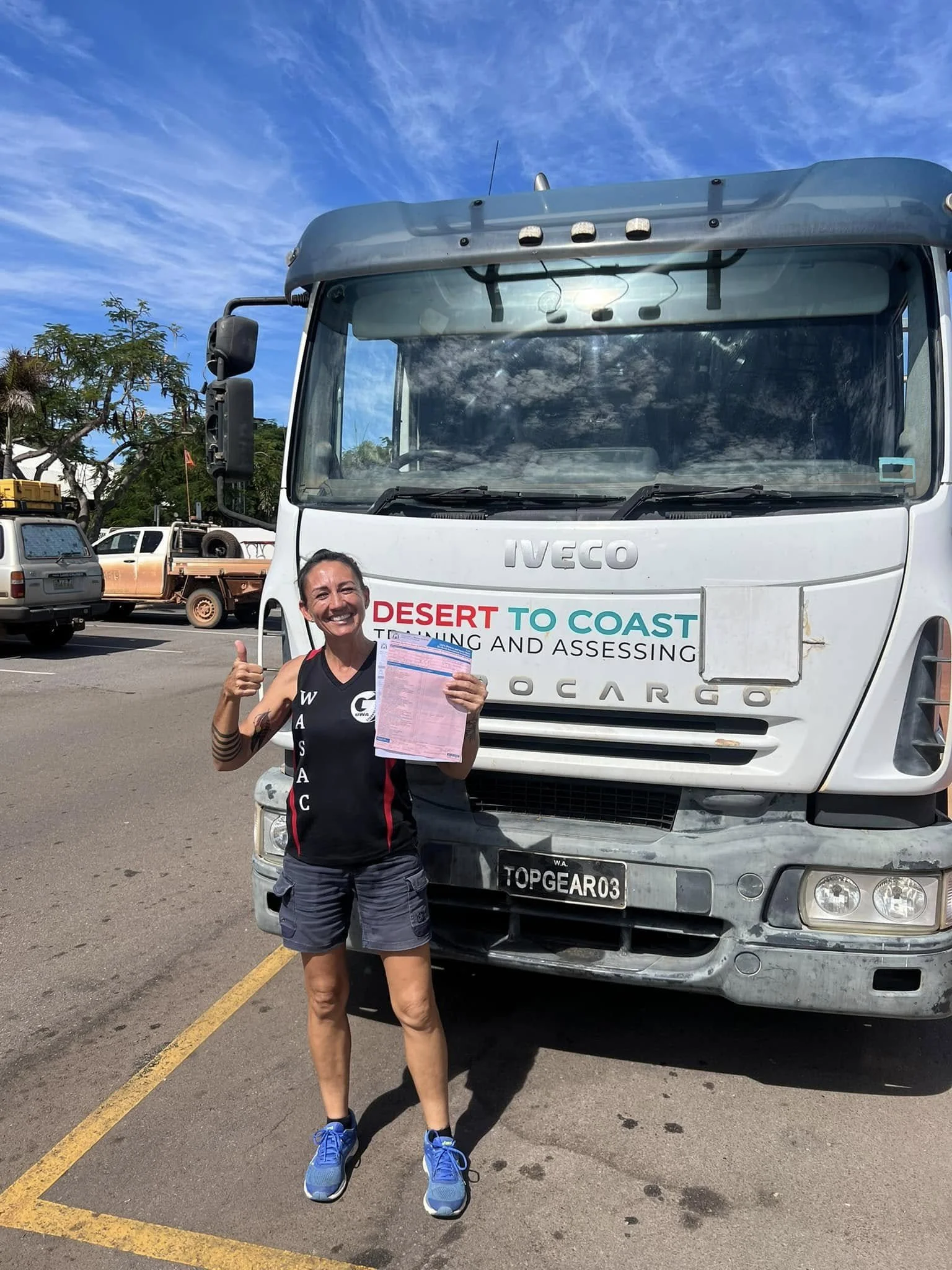 A woman smiling and giving a thumbs up, standing in front of a large white truck with logos and a person holding a paper, in a parking lot with other vehicles and trees, under a partly cloudy sky.