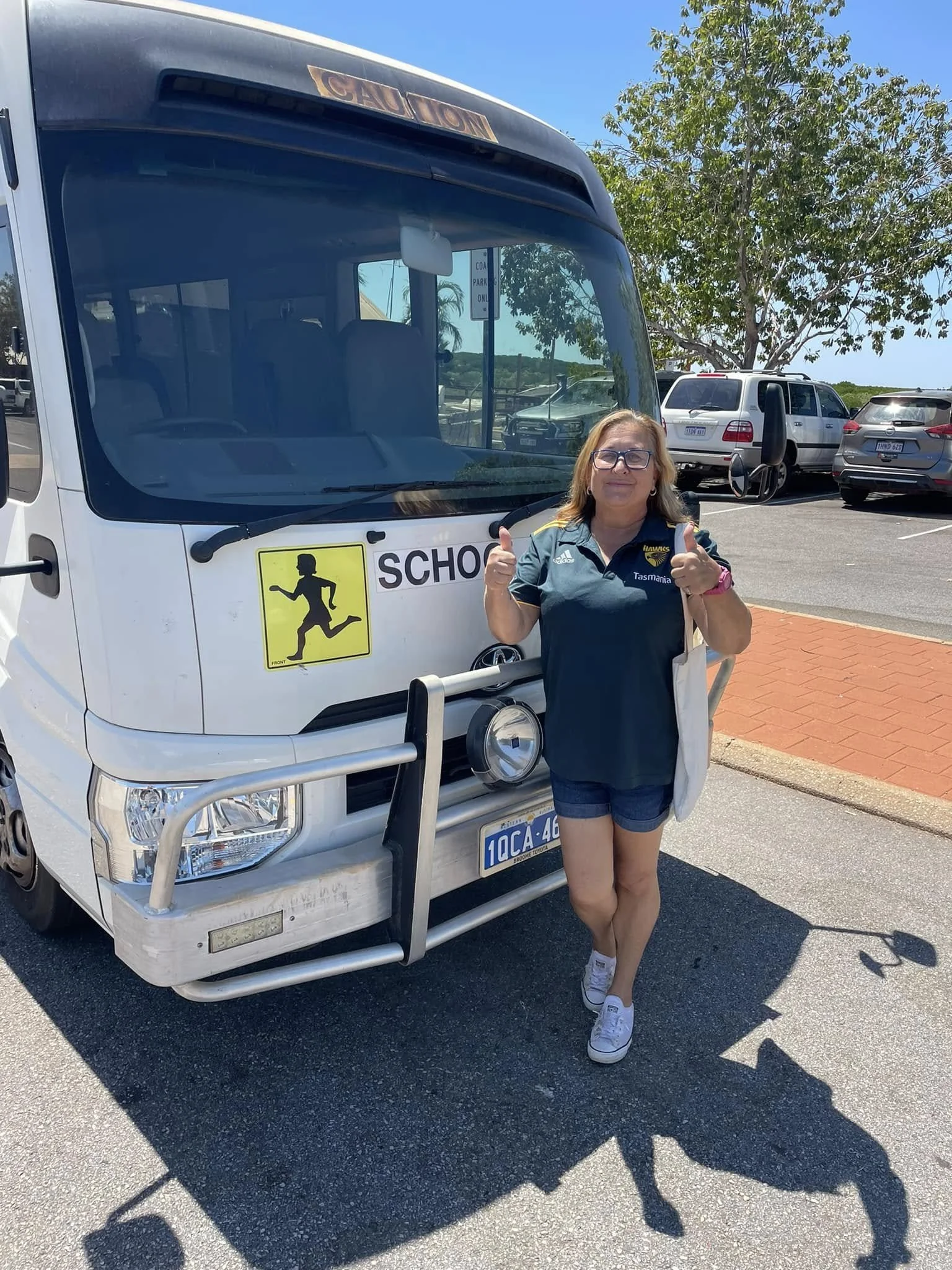 A woman standing next to a white shuttle bus with a school sign on the front, in a parking lot under a tree on a sunny day, giving a thumbs-up gesture.