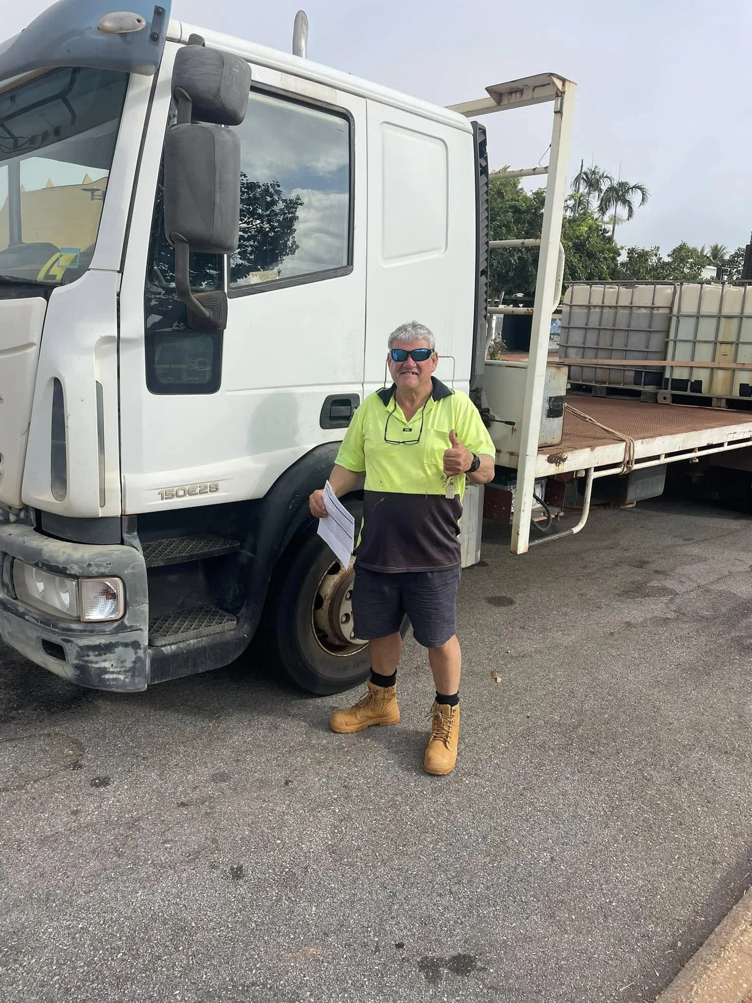 A man in a high-visibility shirt and yellow work boots standing in front of a white flatbed truck, giving a thumbs-up and holding papers, with trees and cloudy sky in the background.