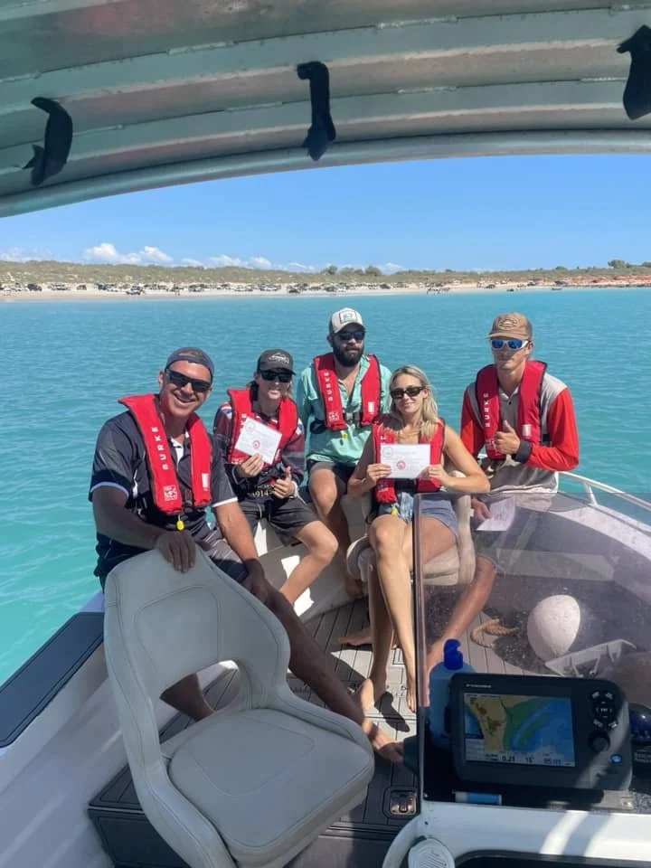 Group of five people on a boat, wearing life jackets, with water and shoreline in the background during daytime.