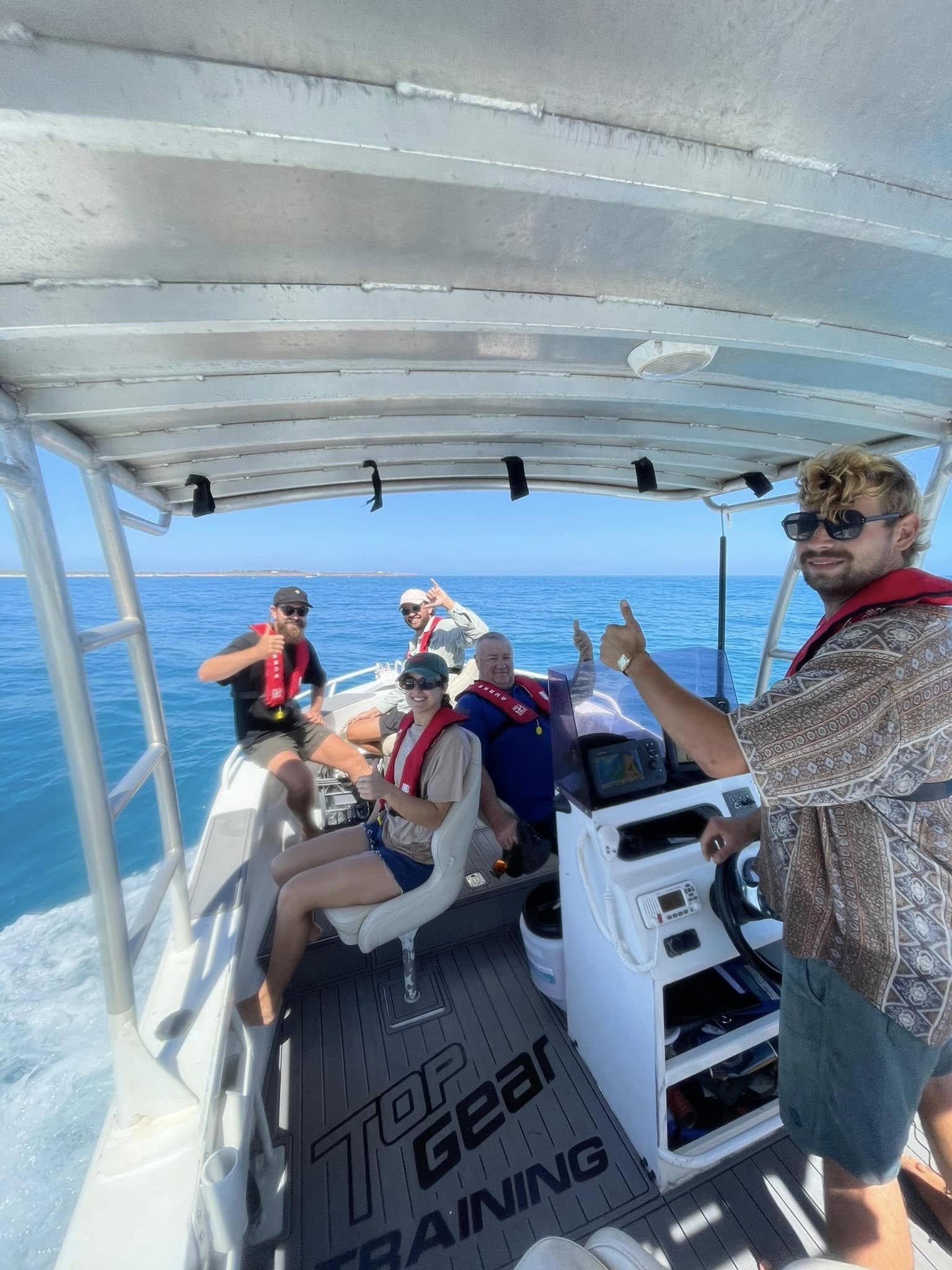 Group of people on a boat enjoying the ocean, some wearing life jackets, with a man in sunglasses and patterned shirt smiling at the camera.