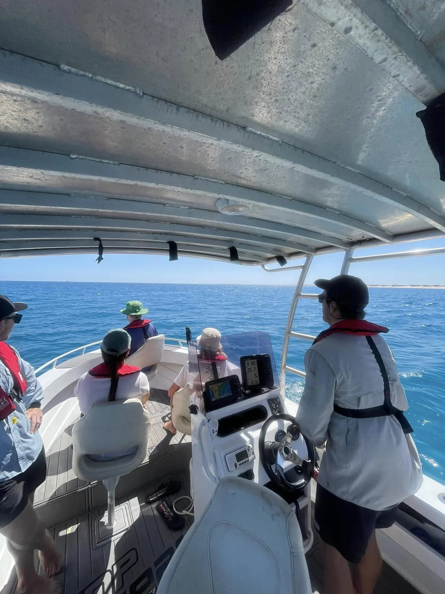 People on a boat ride on the ocean, some are sitting and others are standing near the steering wheel, with a view of the blue sea and sky in the background.