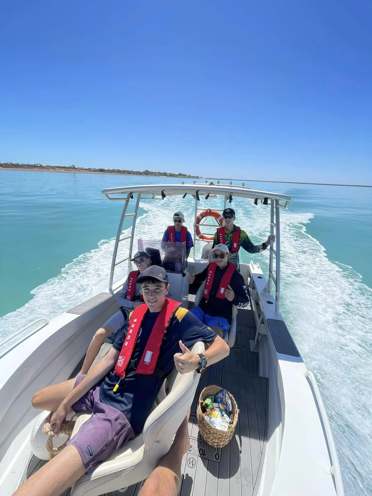 A group of young people on a boat enjoying a sunny day on a calm body of water, with a distant shoreline visible under a clear blue sky.