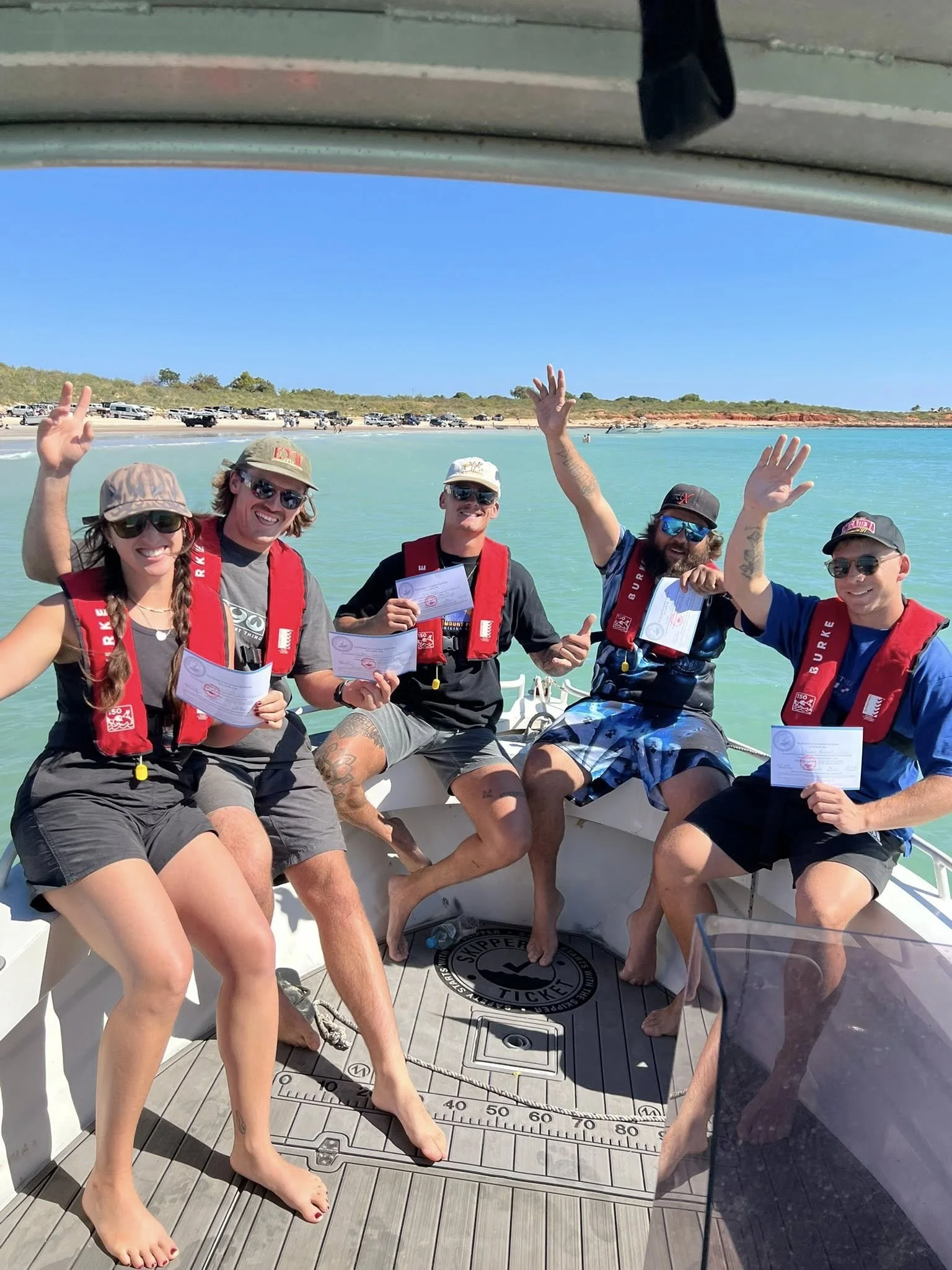 Group of five people on a boat, smile and waving, holding certificates, with a beach and parked cars in the background.