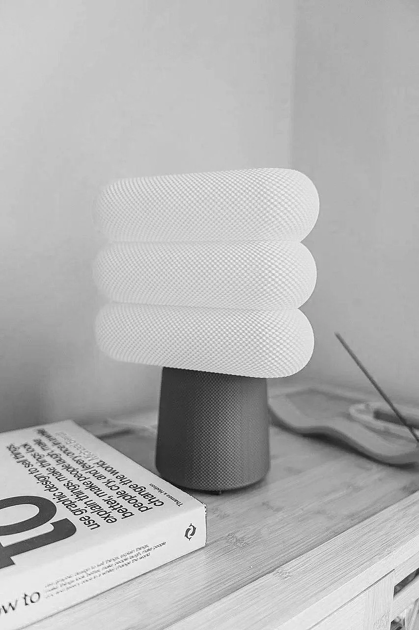 A white, textured, cylindrical lamp with stacked, curved segments sits on a wooden surface next to a book and a small, flat electronic device.