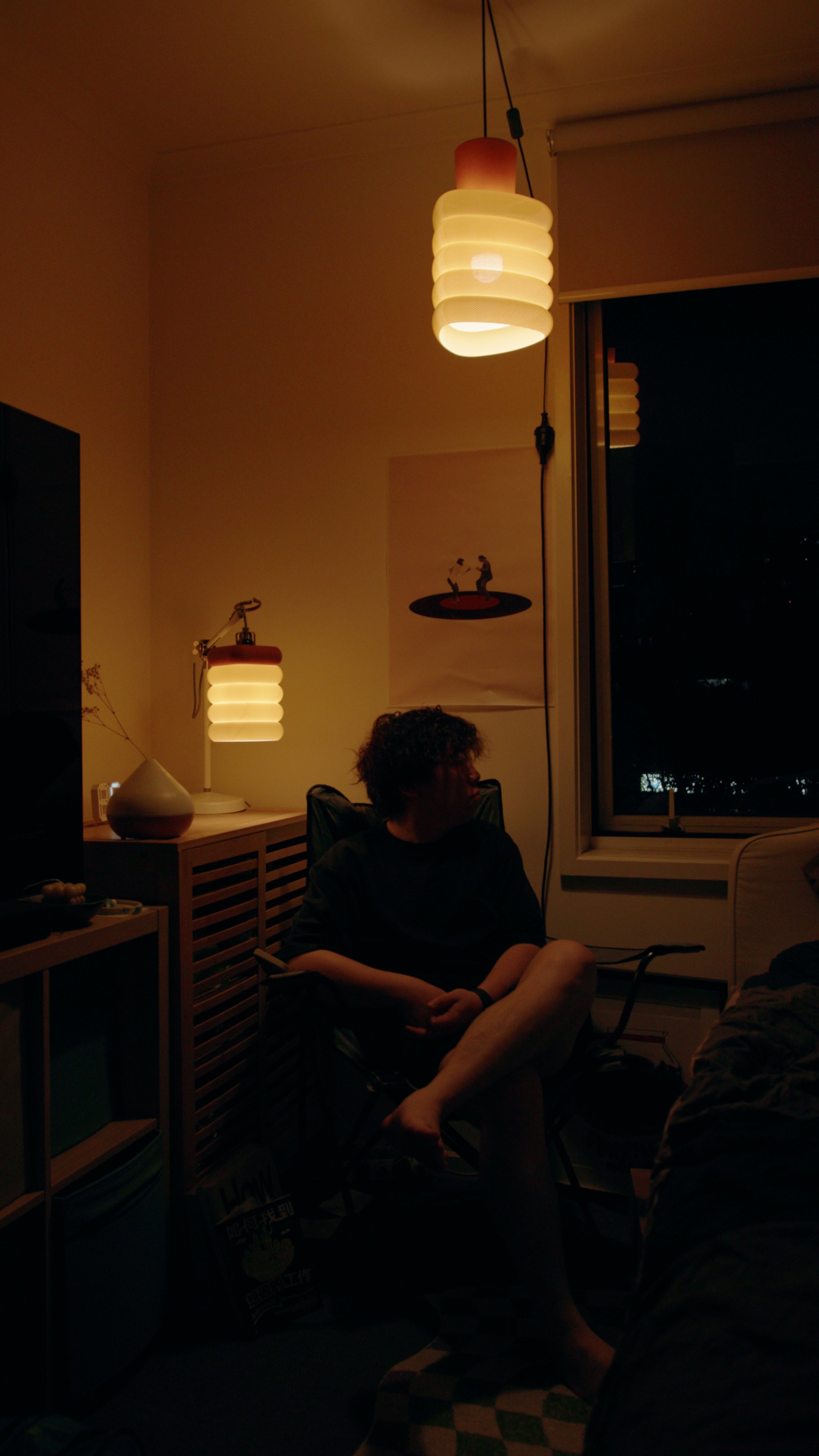 A cozy room illuminated by warm ceiling and desk lamps, with a person sitting on a chair near the window at night.
