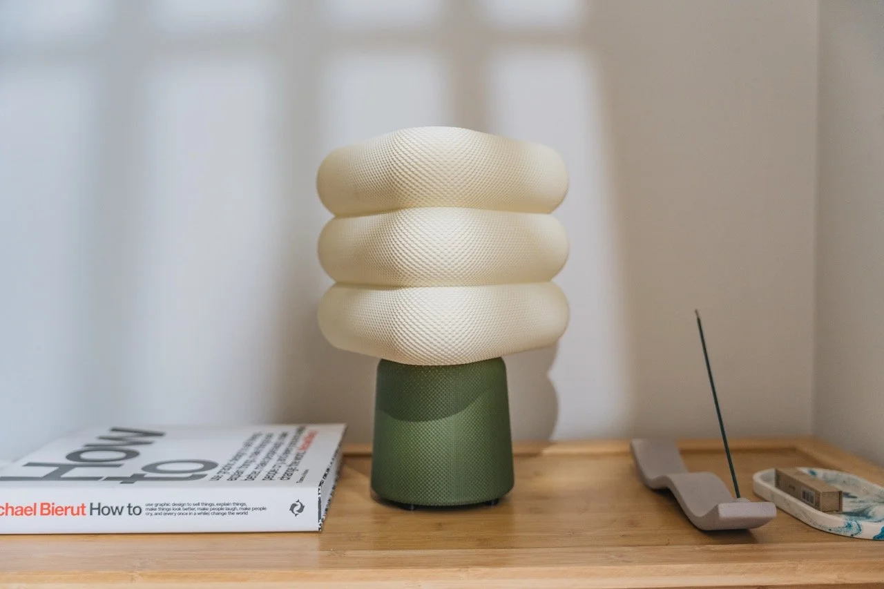 A lamp with a green base and three cushion-like beige lampshades, a magazine, a 3D-printed object, an incense holder with an incense stick, and a stack of paper money on a wooden surface