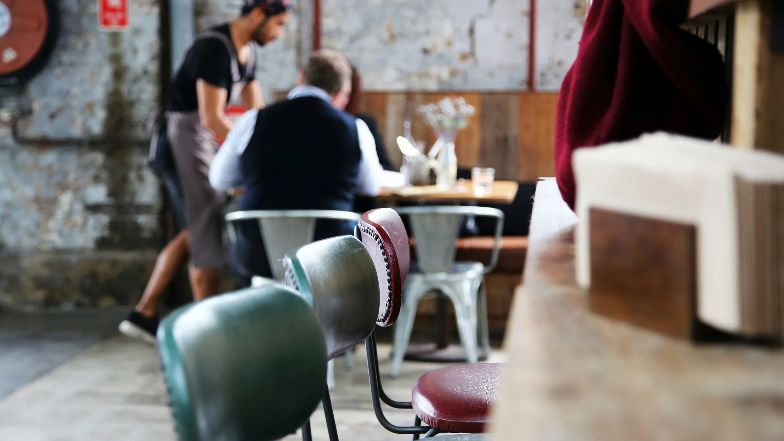 A blurred image showing a restaurant scene with a waitress taking an order from a customer. There are various chairs in the foreground and a rustic wall in the background.