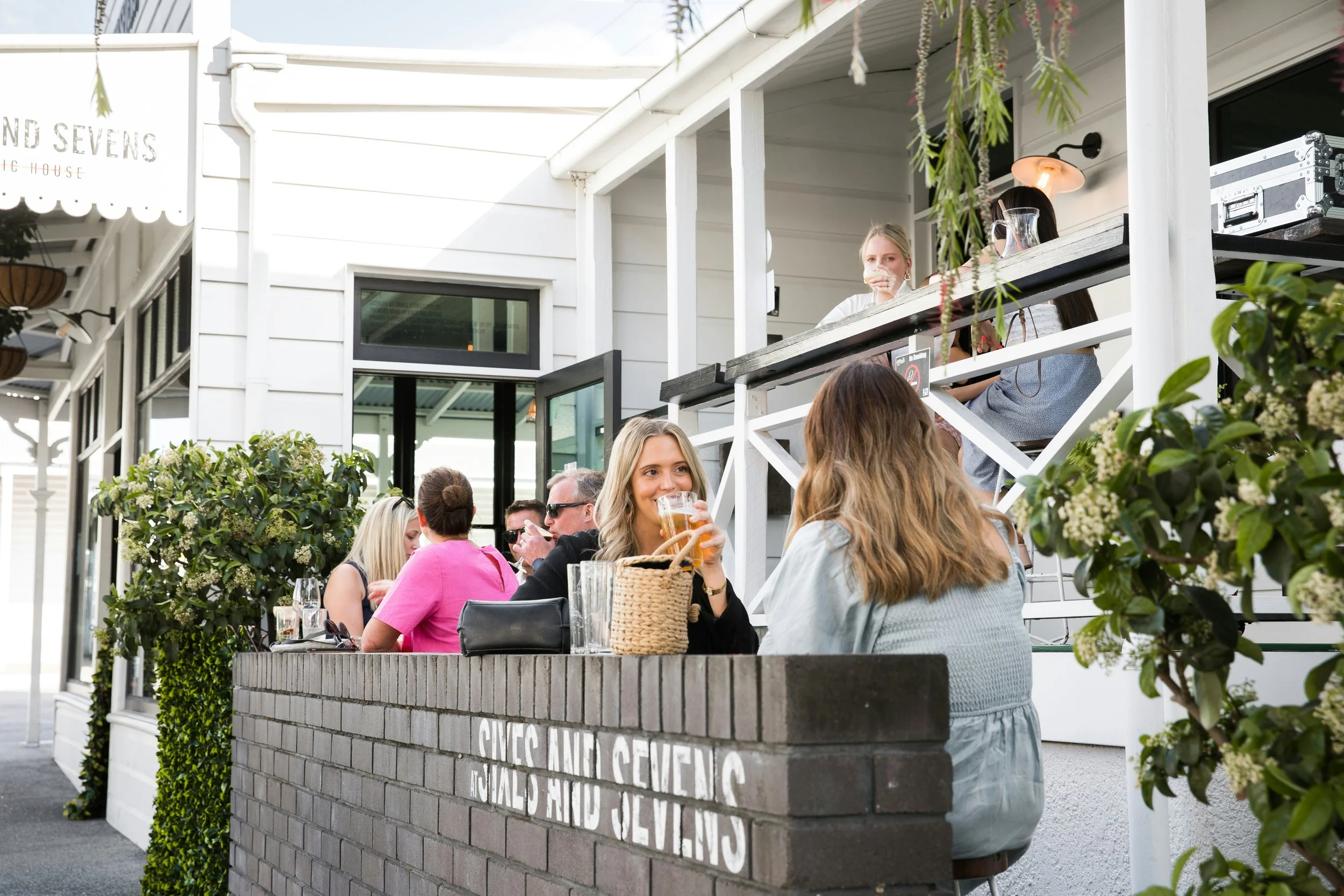 People sitting outside at a cafe or restaurant, enjoying drinks and conversation, with a white building and greenery in the background.