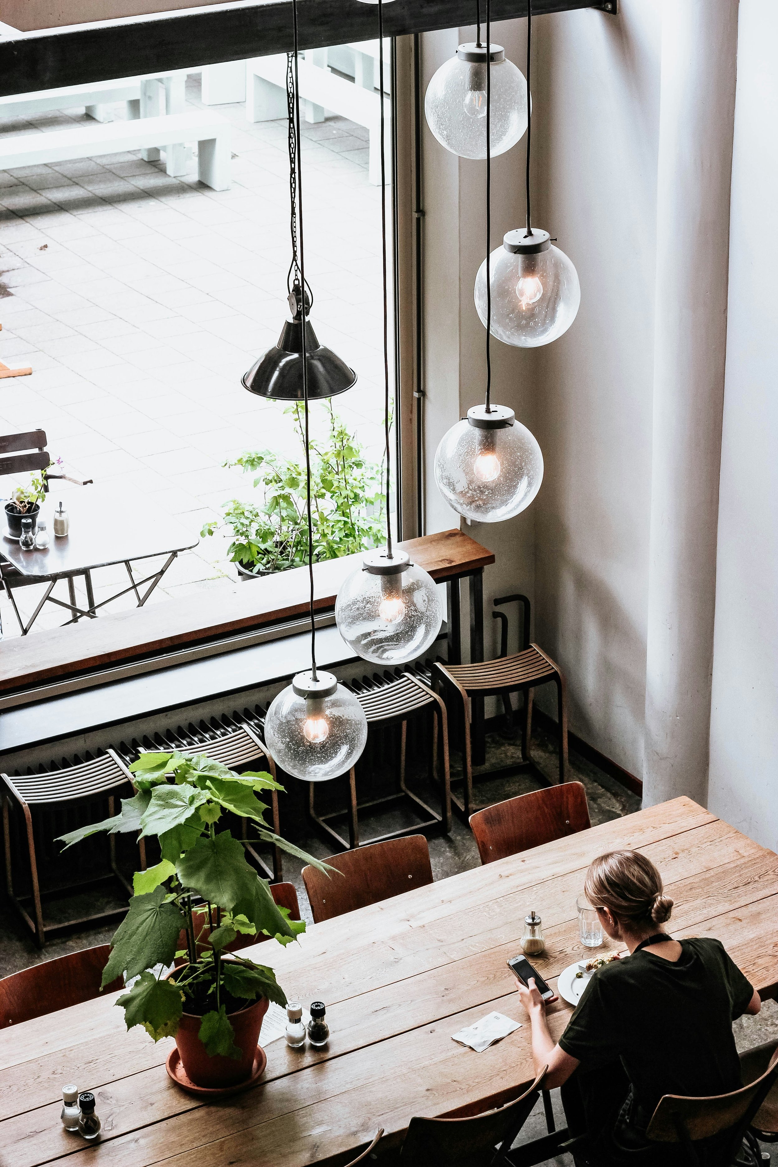 Interior of a café or restaurant with wooden tables, chairs, hanging globe pendant lights, a woman looking at her phone, and greenery outside visible through a large window.