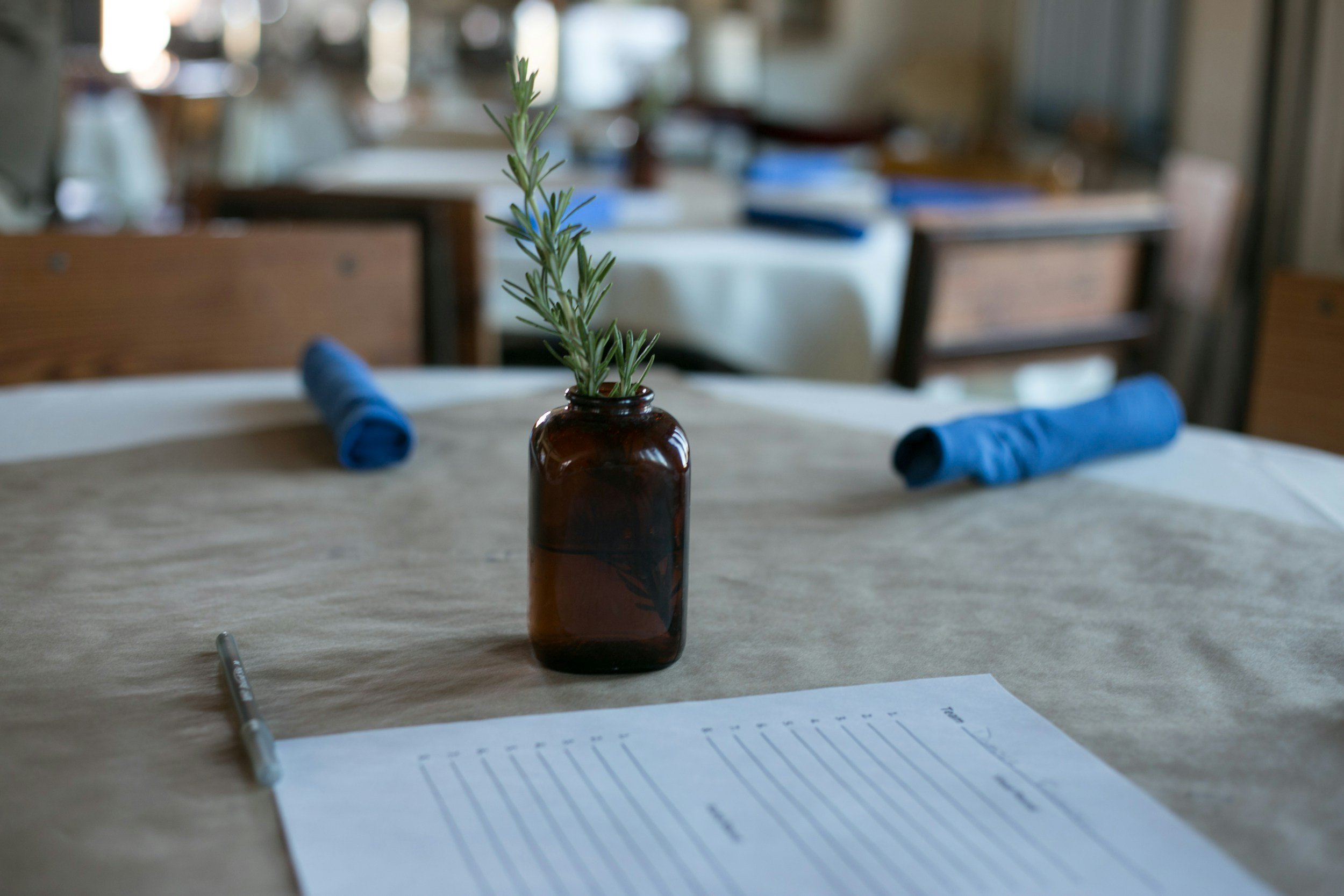 A table with a small plant in a brown bottle, blue rolled cloth napkins, a pen, and a piece of lined paper. In the background, there are chairs and tables, and the room appears to be a restaurant or dining area.