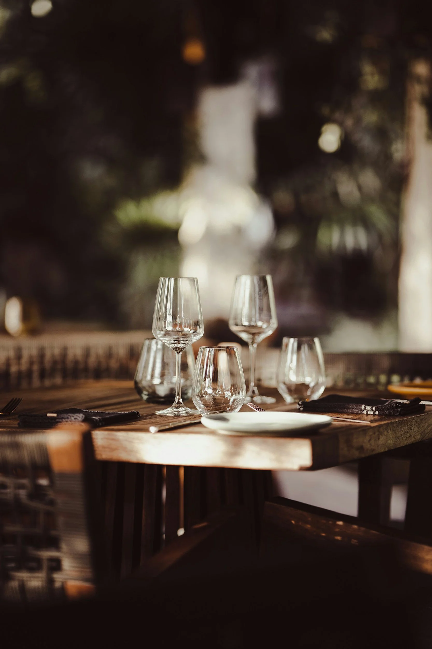 A dining table set with several wine glasses, a white plate, and black napkins, in a warmly lit restaurant with blurred plants or trees in the background.