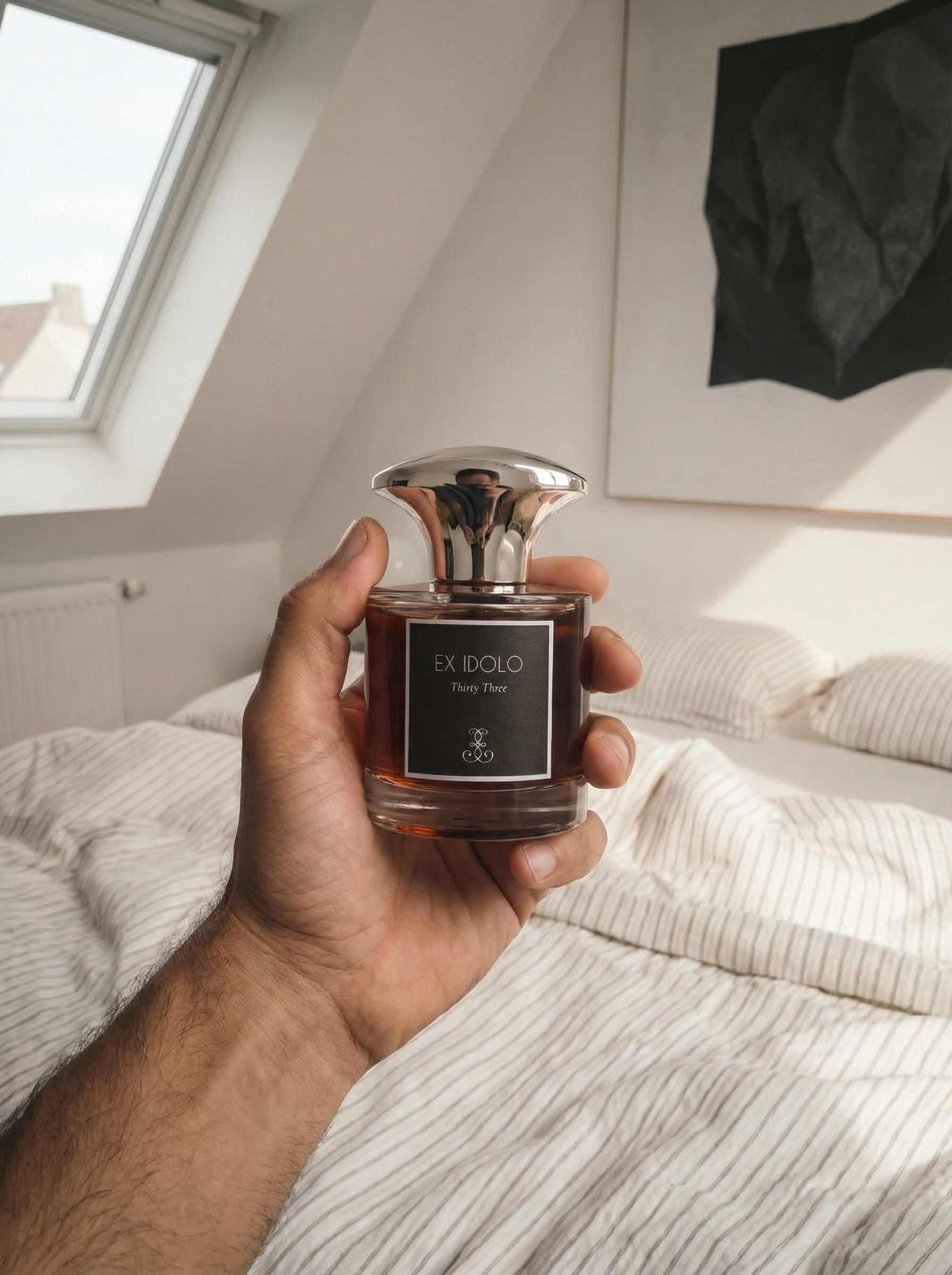 A person's hand holding a perfume bottle labeled 'EX IDOLO Thirty Three' in a bedroom with a window, a black wall art piece, and a bed with striped bedding in the background.