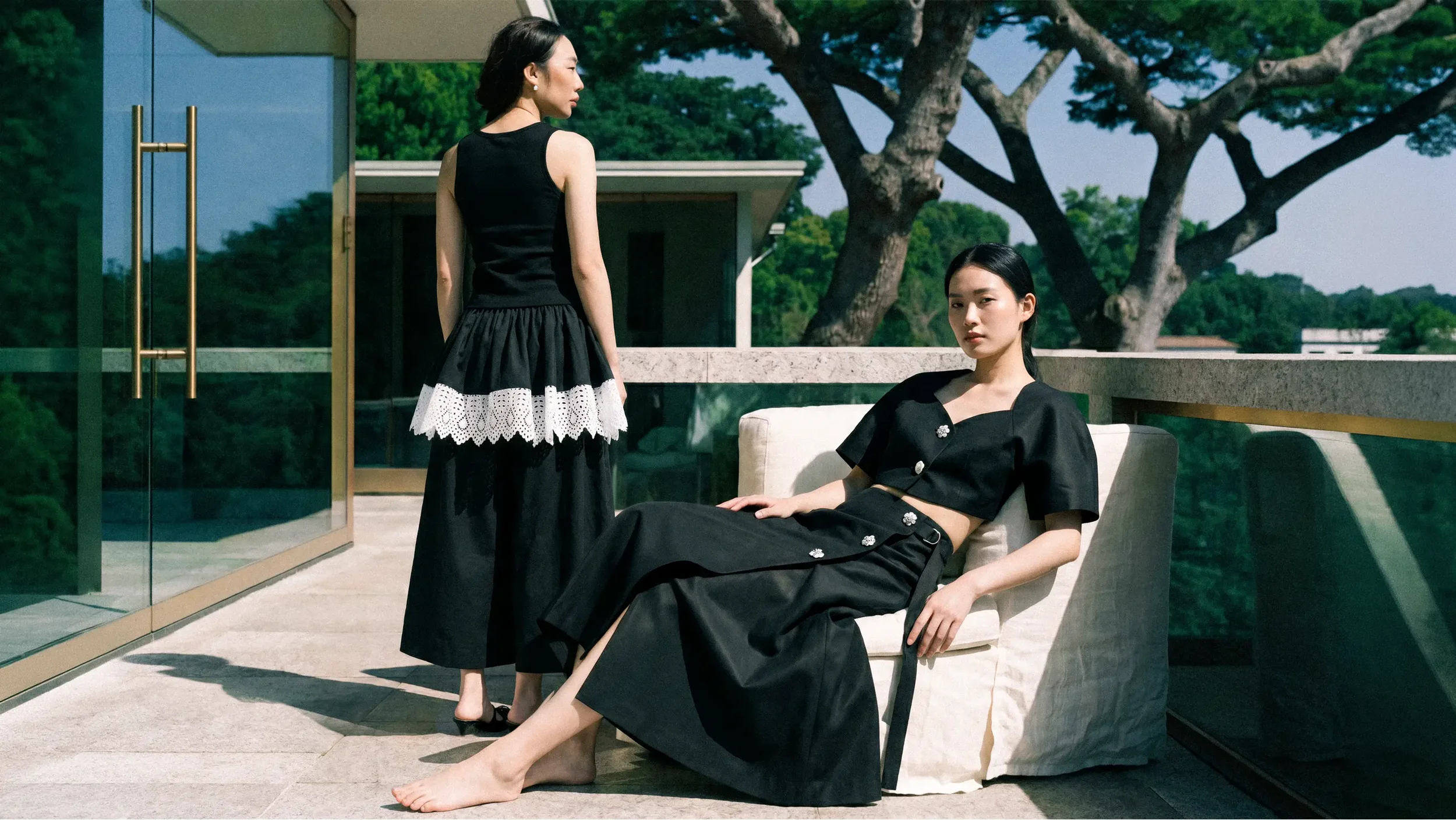 Two women on a modern outdoor patio, one sitting on a white lounge chair, the other standing nearby, with large trees and a clear sky in the background. Both women are dressed in black, with the sitting woman in a black dress and the standing woman in a sleeveless black top and black skirt with a white lace trim.