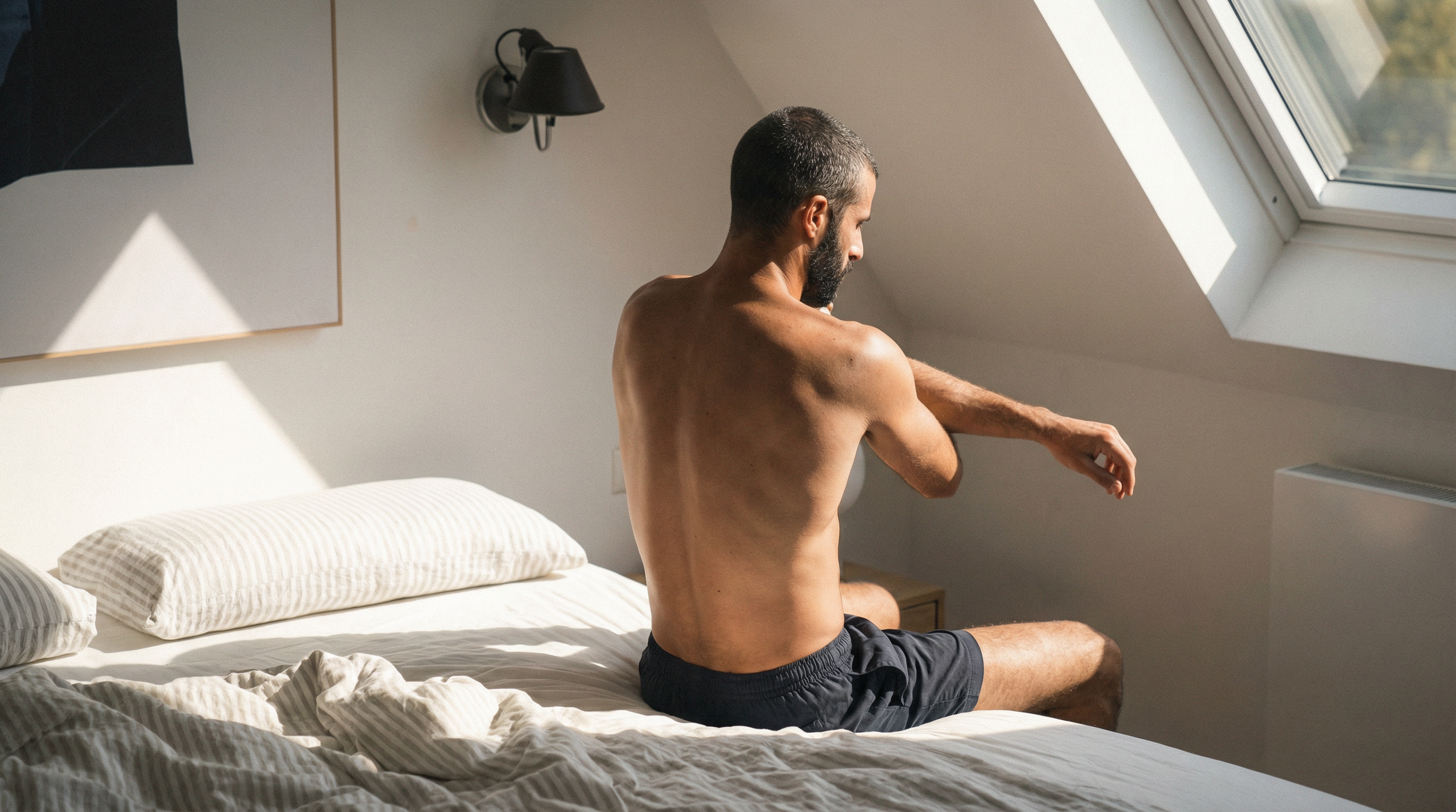A shirtless man with dark hair and beard stretching while sitting on a bed in a sunlit room with a slanted ceiling and a large window.