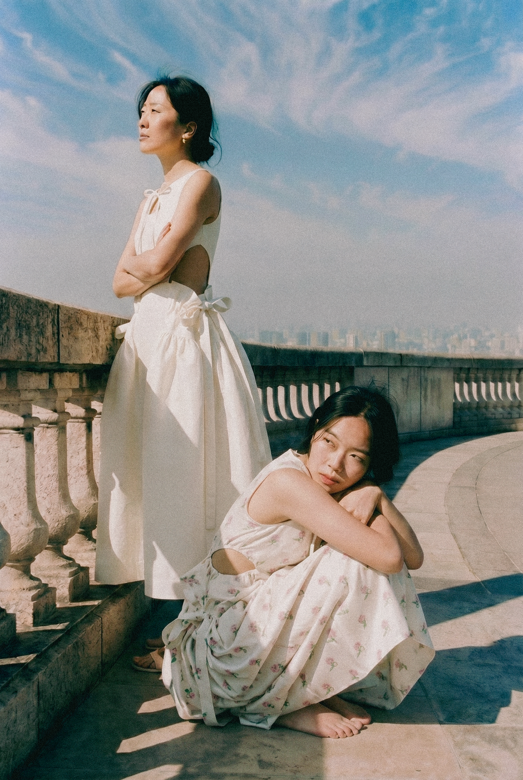 Two women are sitting outdoors on a stone ledge with a city skyline in the background. One woman is standing with arms crossed, wearing a white sleeveless dress, looking to the side. The other woman is sitting with knees drawn up, resting her head on her arms, wearing a light-colored floral dress, gazing to the side as well.